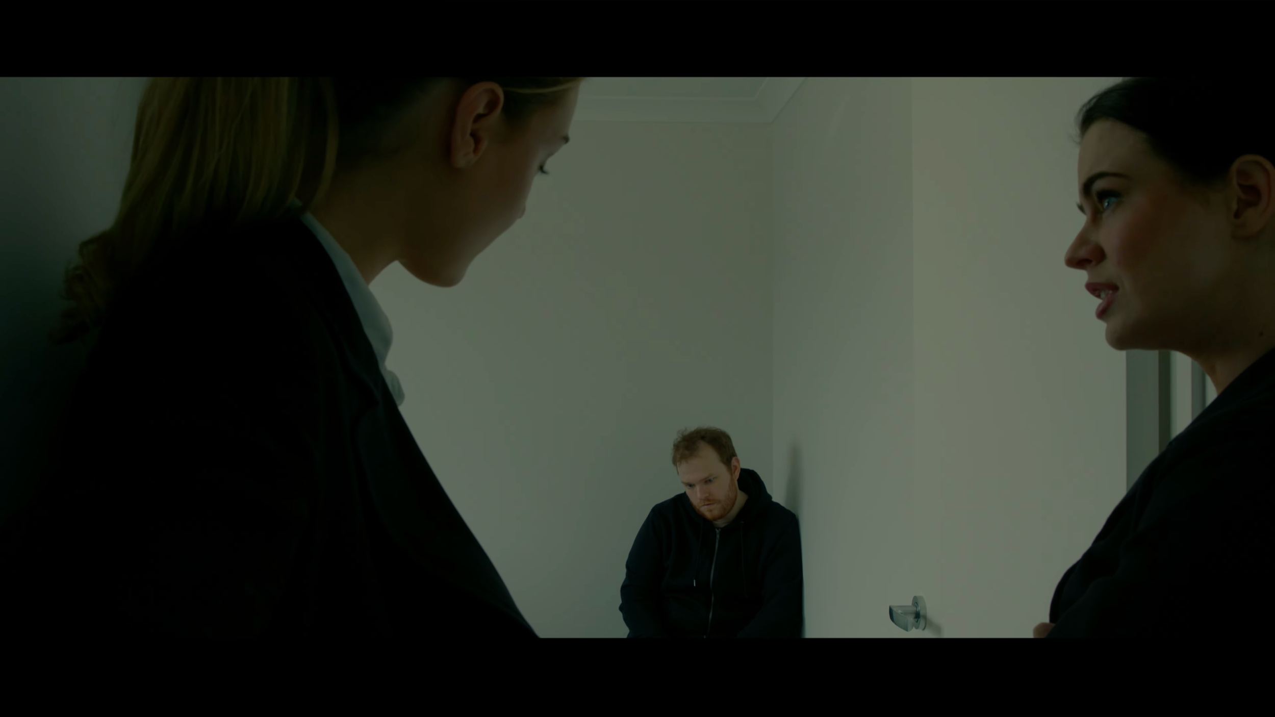 Two women in a confrontation in a room, with a man sitting on a chair in the background watching.