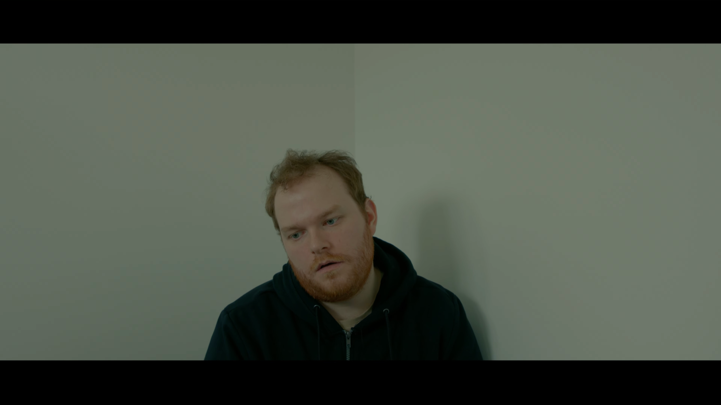 A young man with reddish hair and a beard sitting against a plain white wall, wearing a black hoodie, looking down with a serious expression.