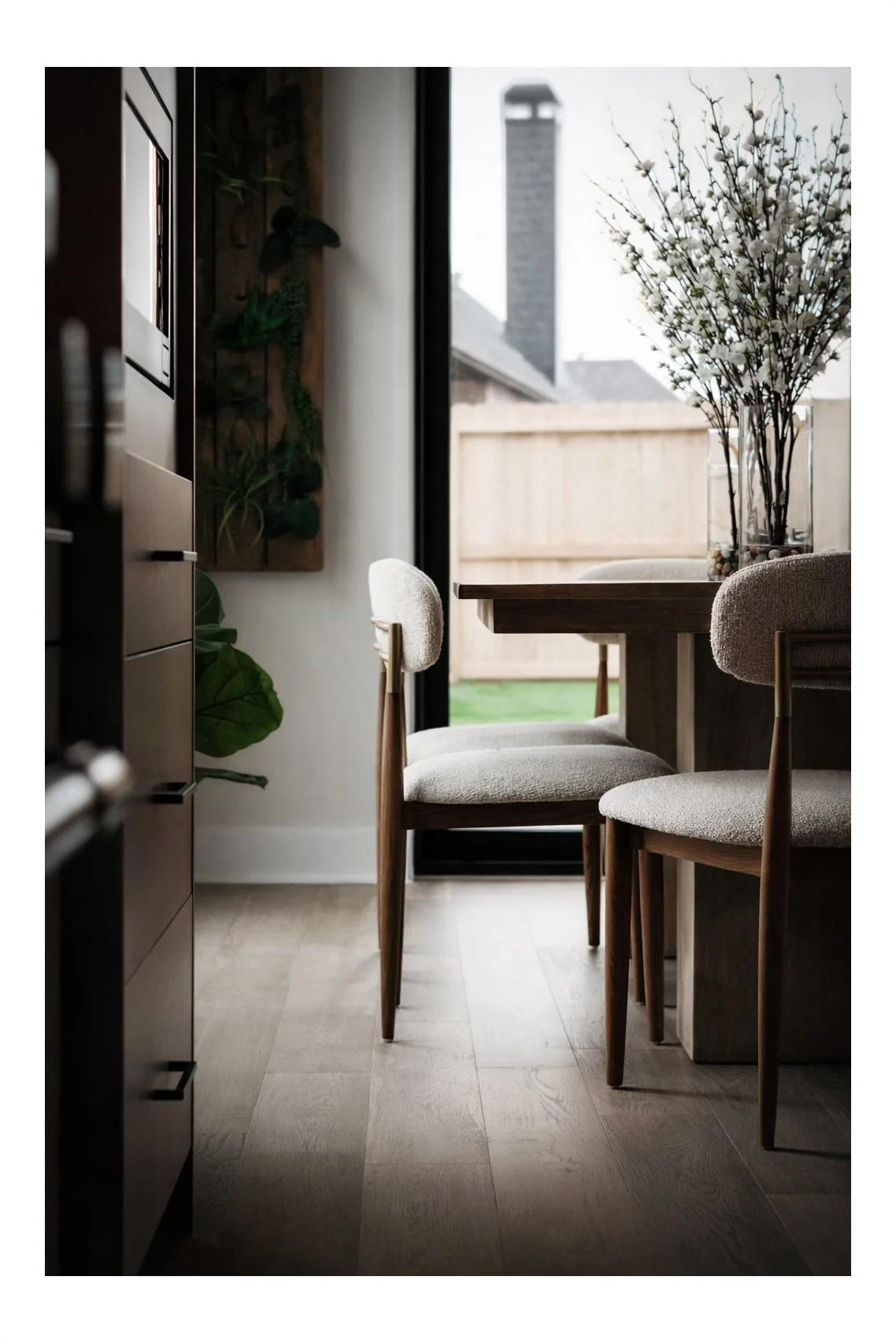 A cozy dining area with a wooden table, two cushioned chairs, a large glass vase of white flowers, and a view of a backyard with a wooden fence and a chimney