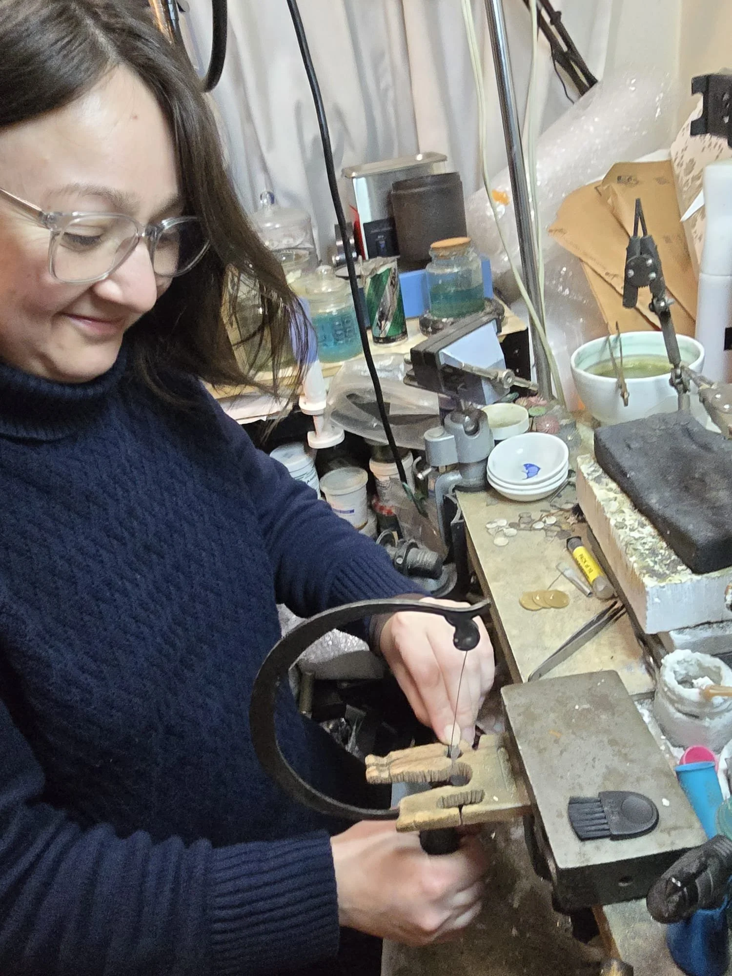 Aliza Souleyeva working at a jeweler's workbench, holding a small object with a bow drill, surrounded by jewelry-making tools and supplies.