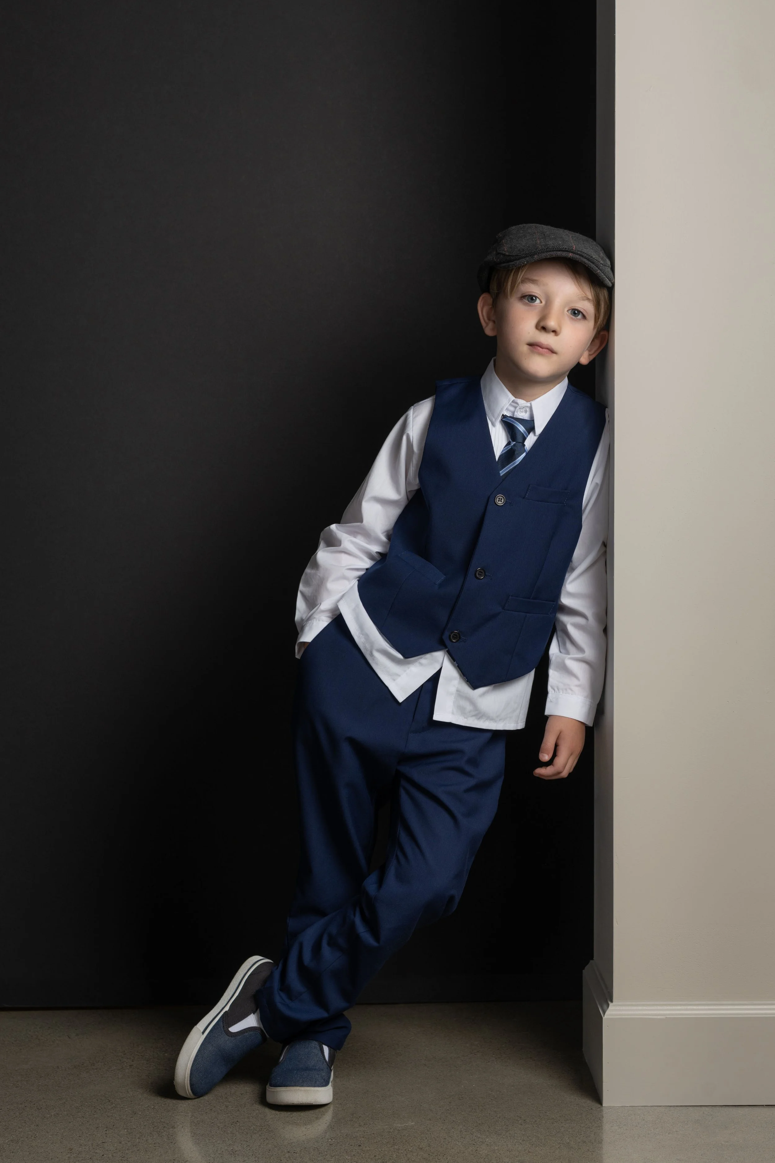 Boy leaning against a pillar, wearing dark blue dress pants and vest with untucked white shirt, tie, casual shoes, and old fashioned cap.