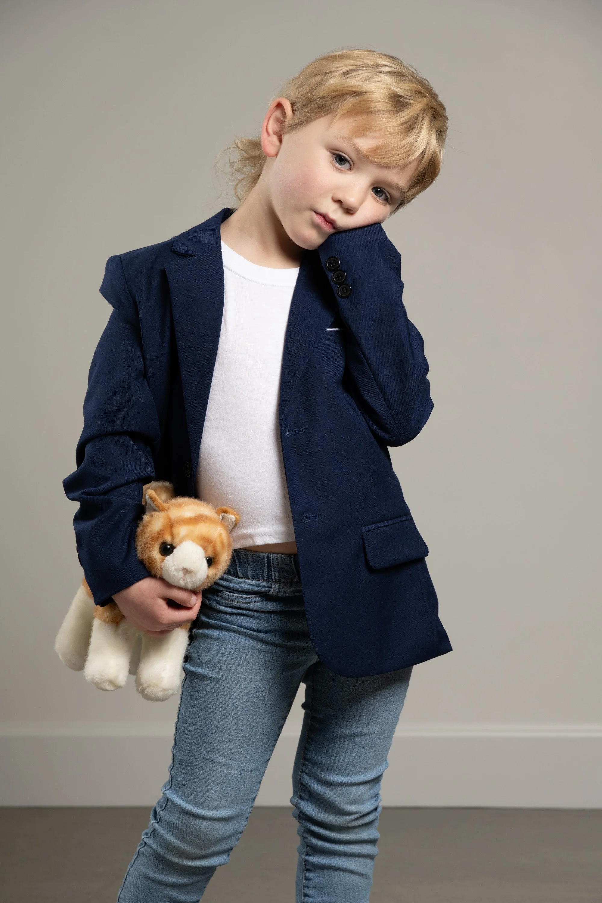 Blond-haired boy wearing jeans and tee shirt with dark blue blazer over the top, standing awkwardly and holding a stuffed cat at his hip.