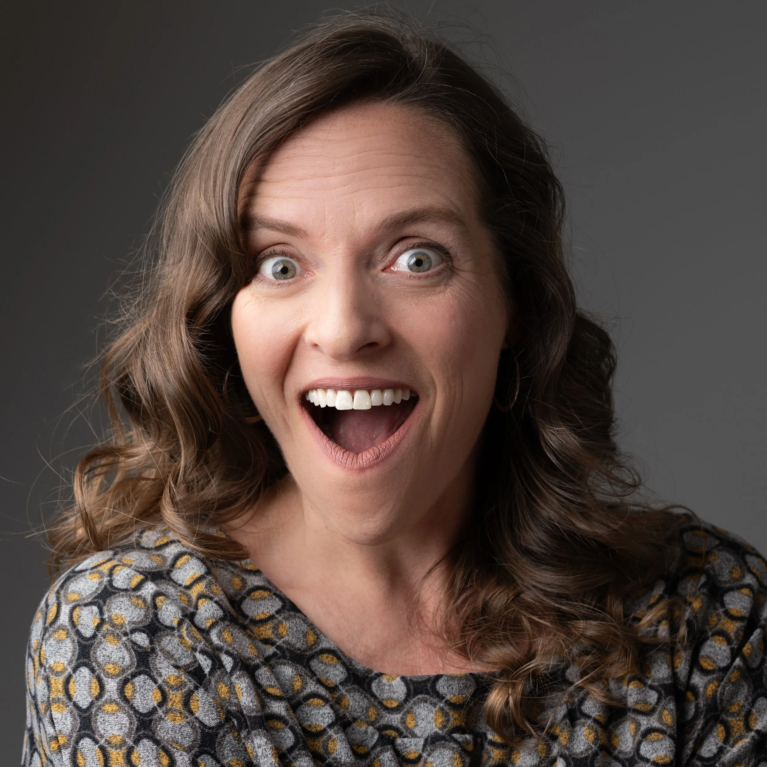 Close-up portrait of a woman with wavy brown hair, green eyes, and hoop earrings, wearing a patterned top and looking directly at the camera.