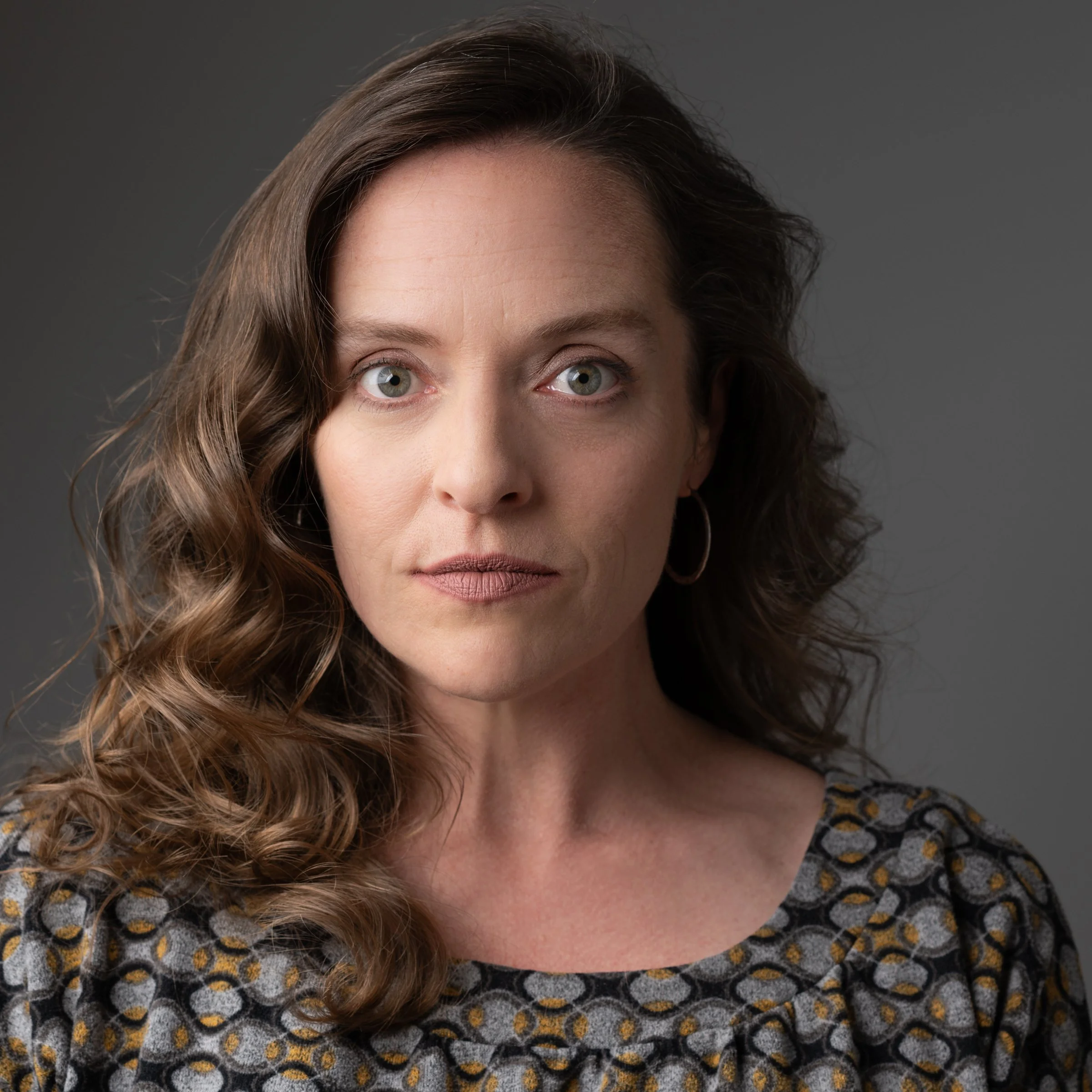 Close-up portrait of a woman with wavy brown hair, green eyes, and hoop earrings, wearing a patterned top and looking directly at the camera.