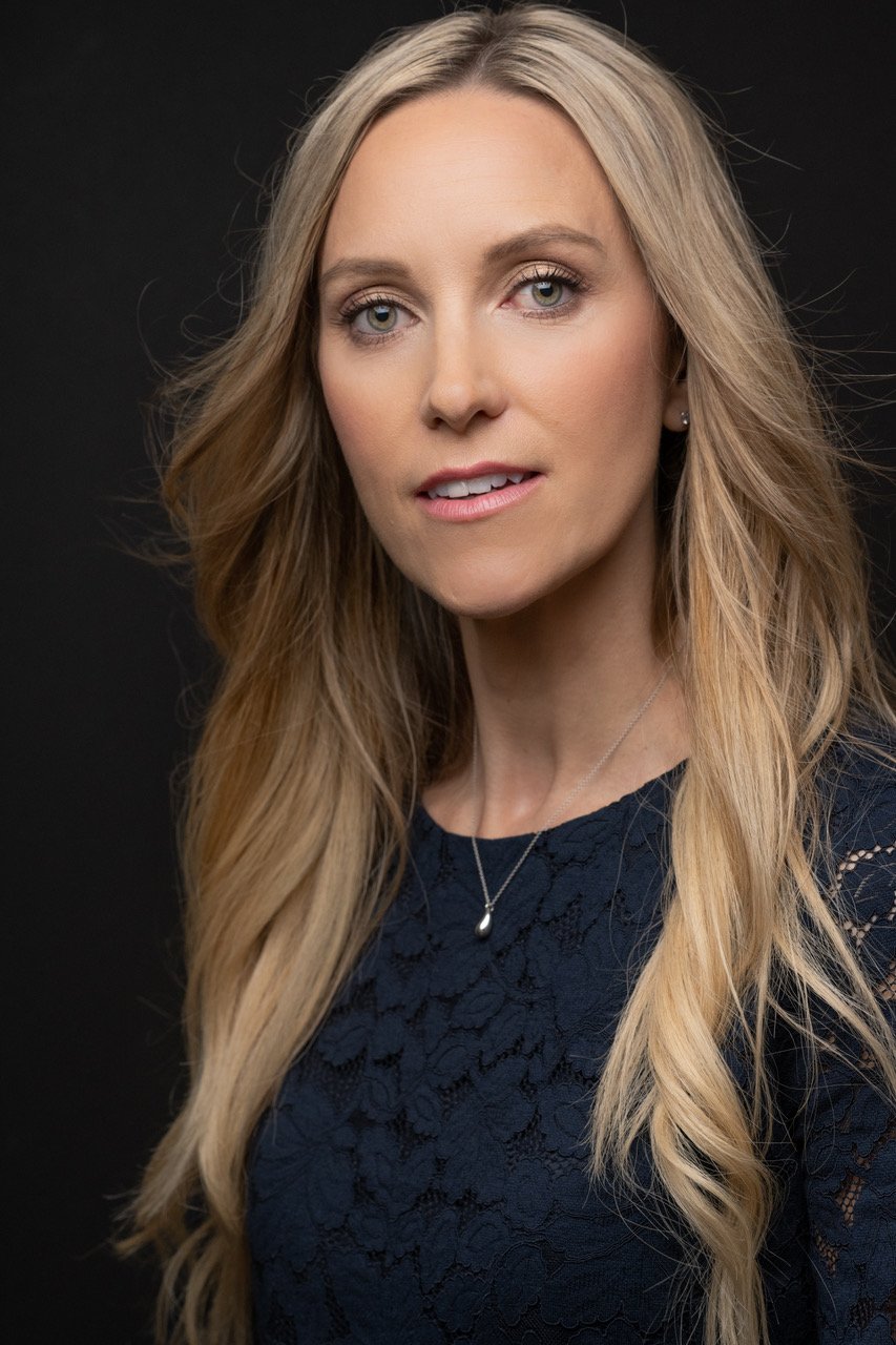 A smiling woman with long blonde hair sitting in a white chair inside a bright room with white curtains and a small table with a flower arrangement.