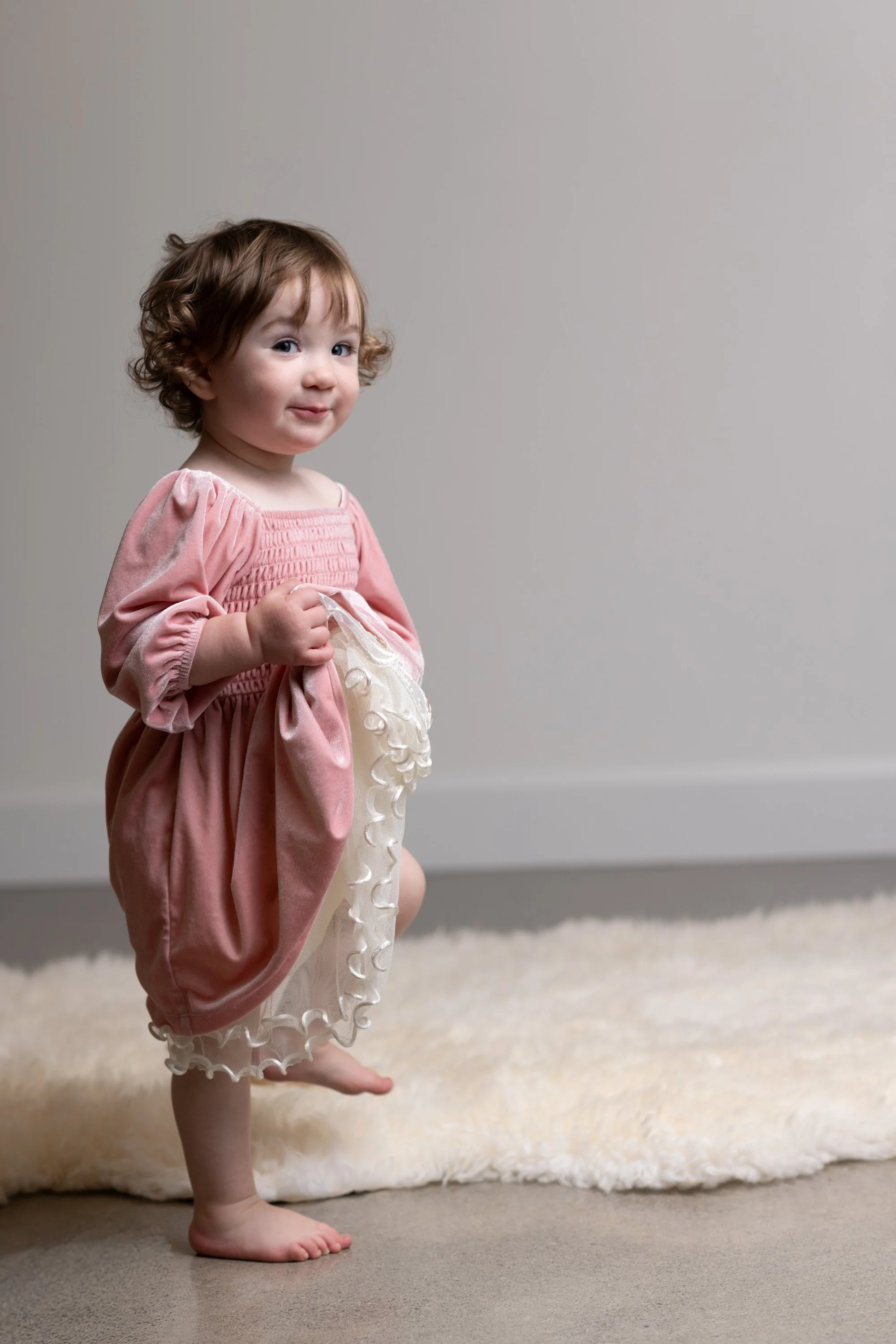 Toddler girl in a pink velvet dress with petticoat, holding the skirt and smirking.