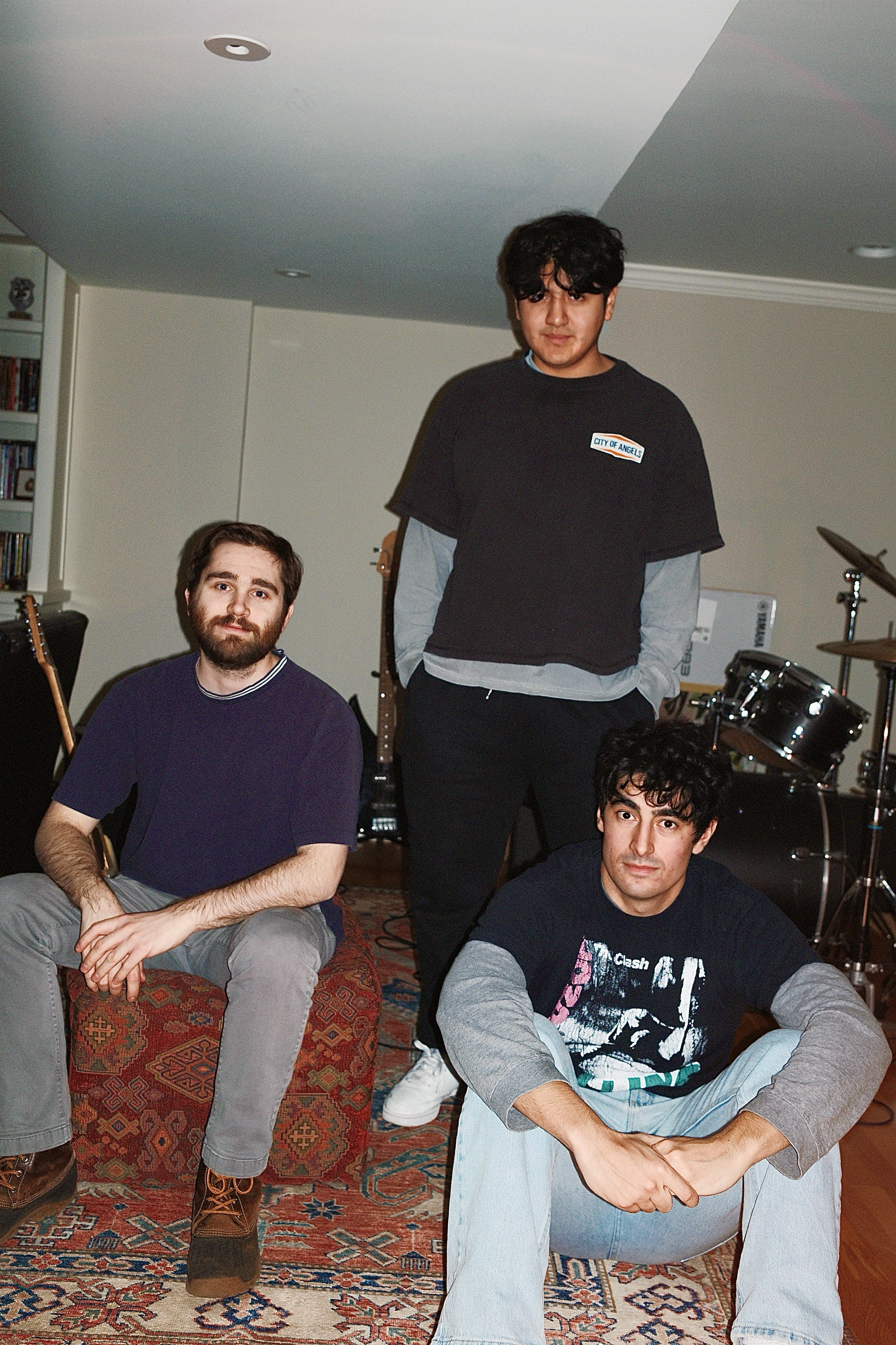 A young man with dark, curly hair and light skin standing next to a stone wall. He is wearing a dark overcoat, a patterned shirt, black pants, and white shoes. He is looking at the camera with a neutral expression, with hand in pocket, next to a pathway with fallen leaves.