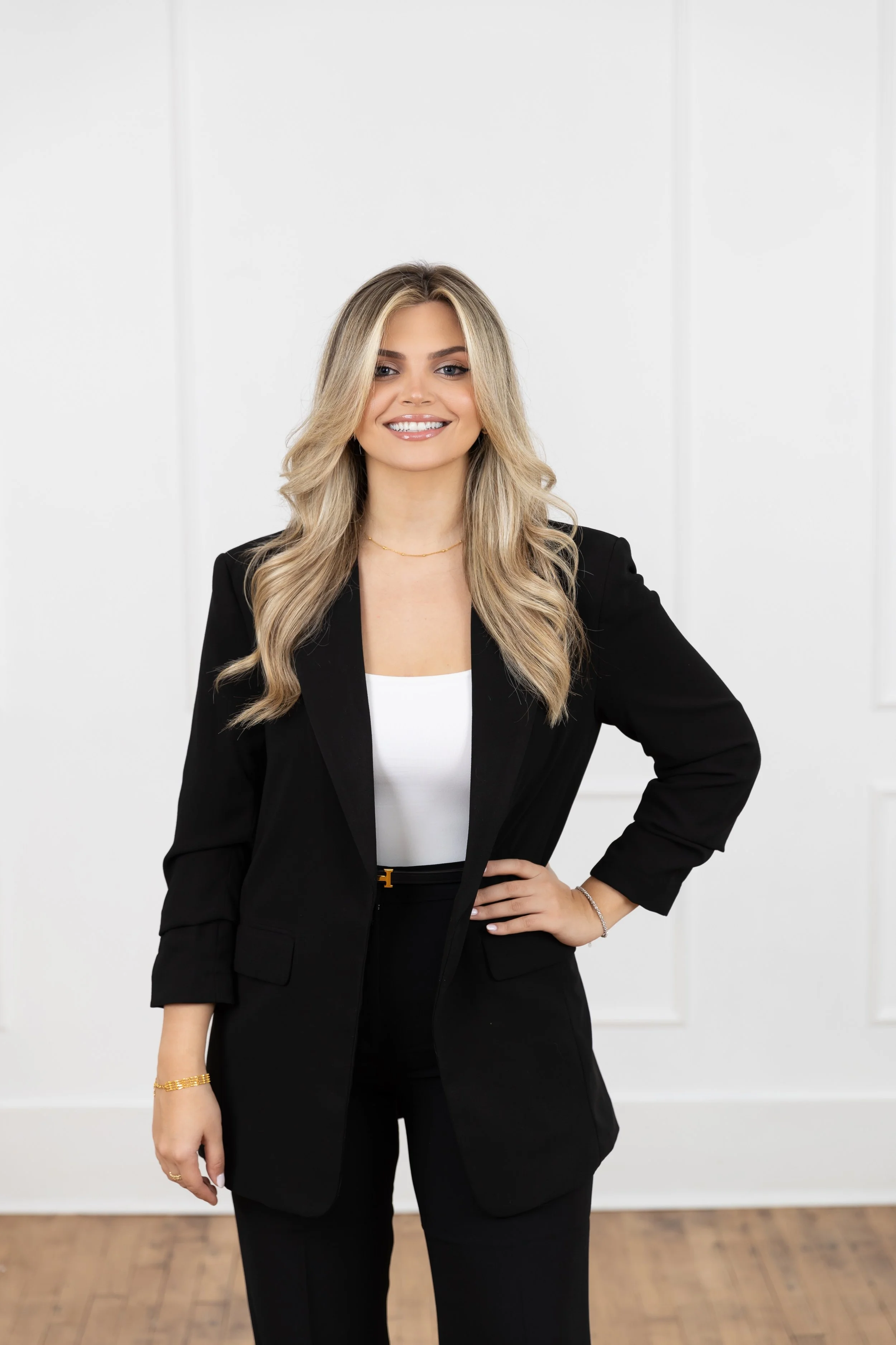 A woman with long blonde hair wearing a black blazer and trousers, a white top, standing with one hand on her hip in a bright, minimalist room.