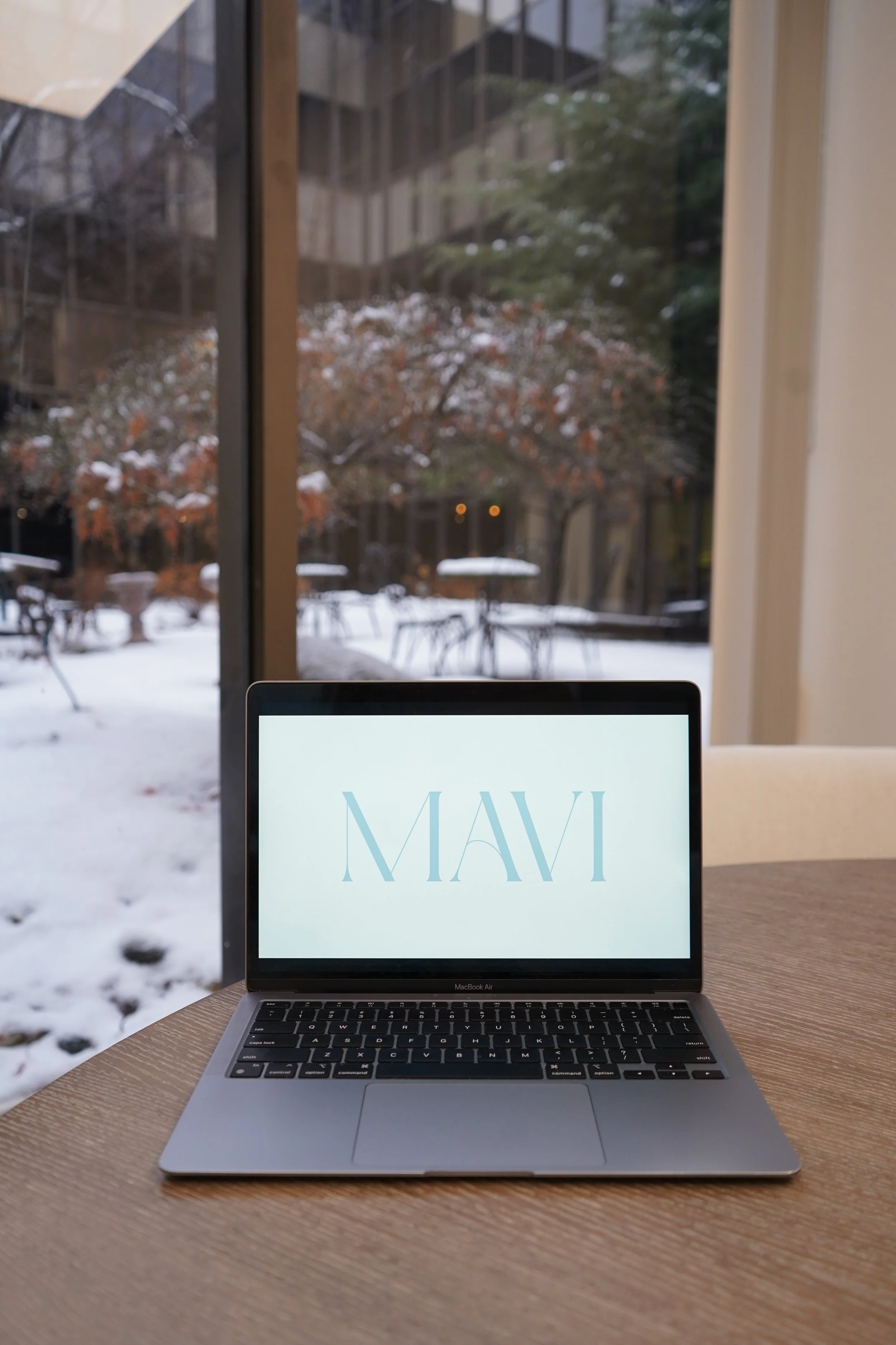 A MacBook Air laptop on a wooden table displaying the word 'MAVI' on its screen, with a snowy outdoor scene and patio furniture visible through a large glass window in the background.