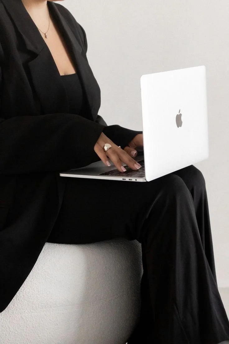 A woman wearing a black suit and jewelry sitting on a white surface and working on an Apple MacBook laptop.