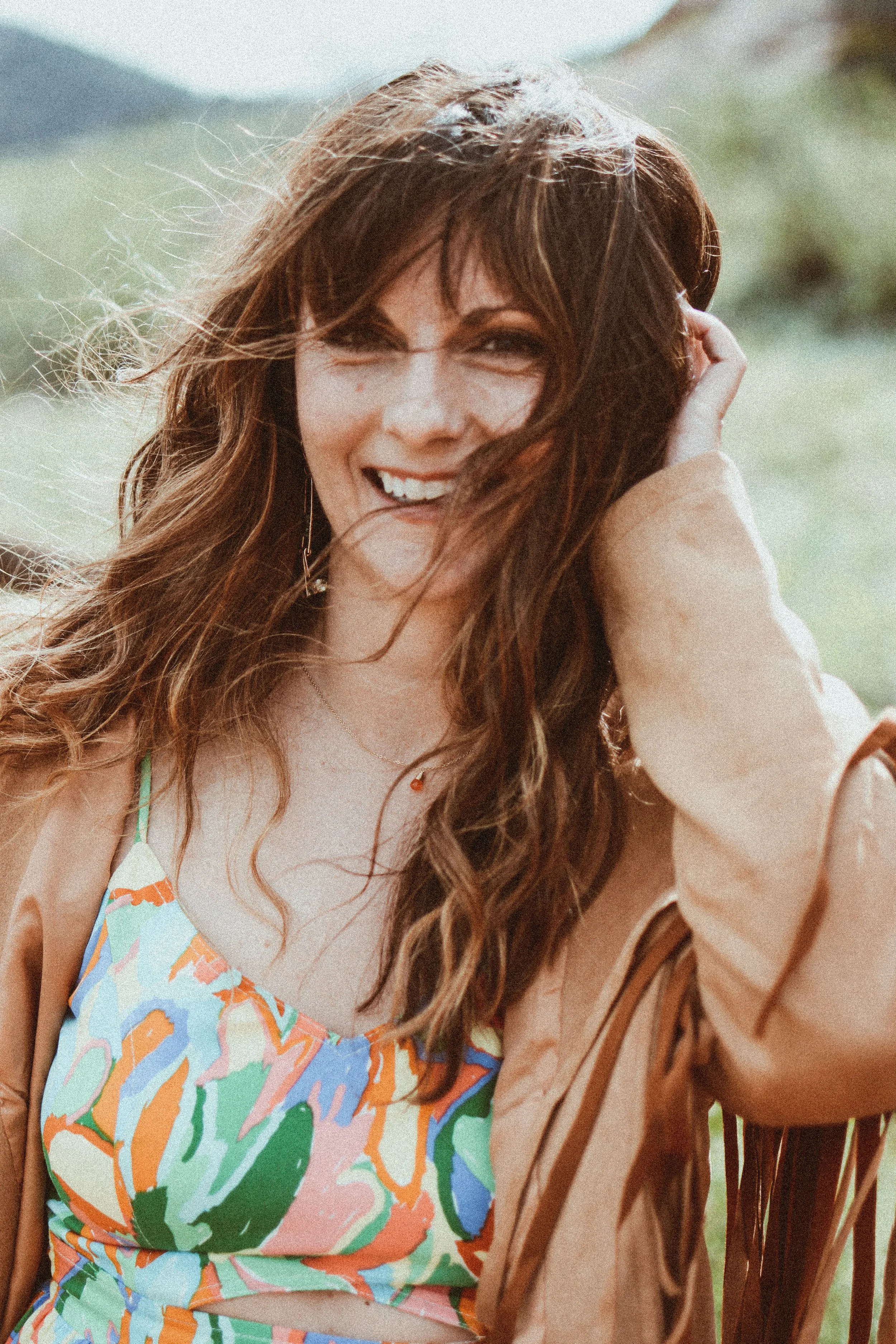A woman with long, wavy, red hair smiling outdoors in a natural setting with mountains in the background, wearing a colorful sleeveless top and a tan jacket.