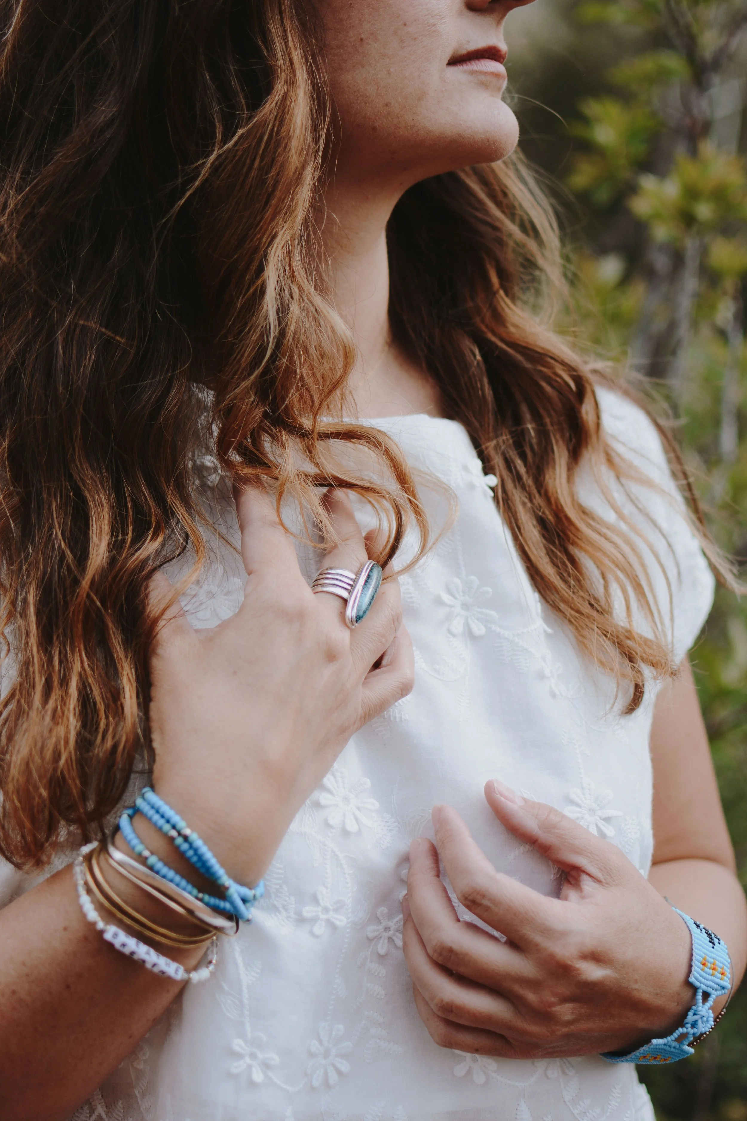 Close-up of a woman with wavy auburn hair wearing a white embroidered dress, with her left hand resting on her chest and her right hand touching her shoulder, adorned with rings, bracelets, and beaded accessories, outdoors with blurred greenery in the background.