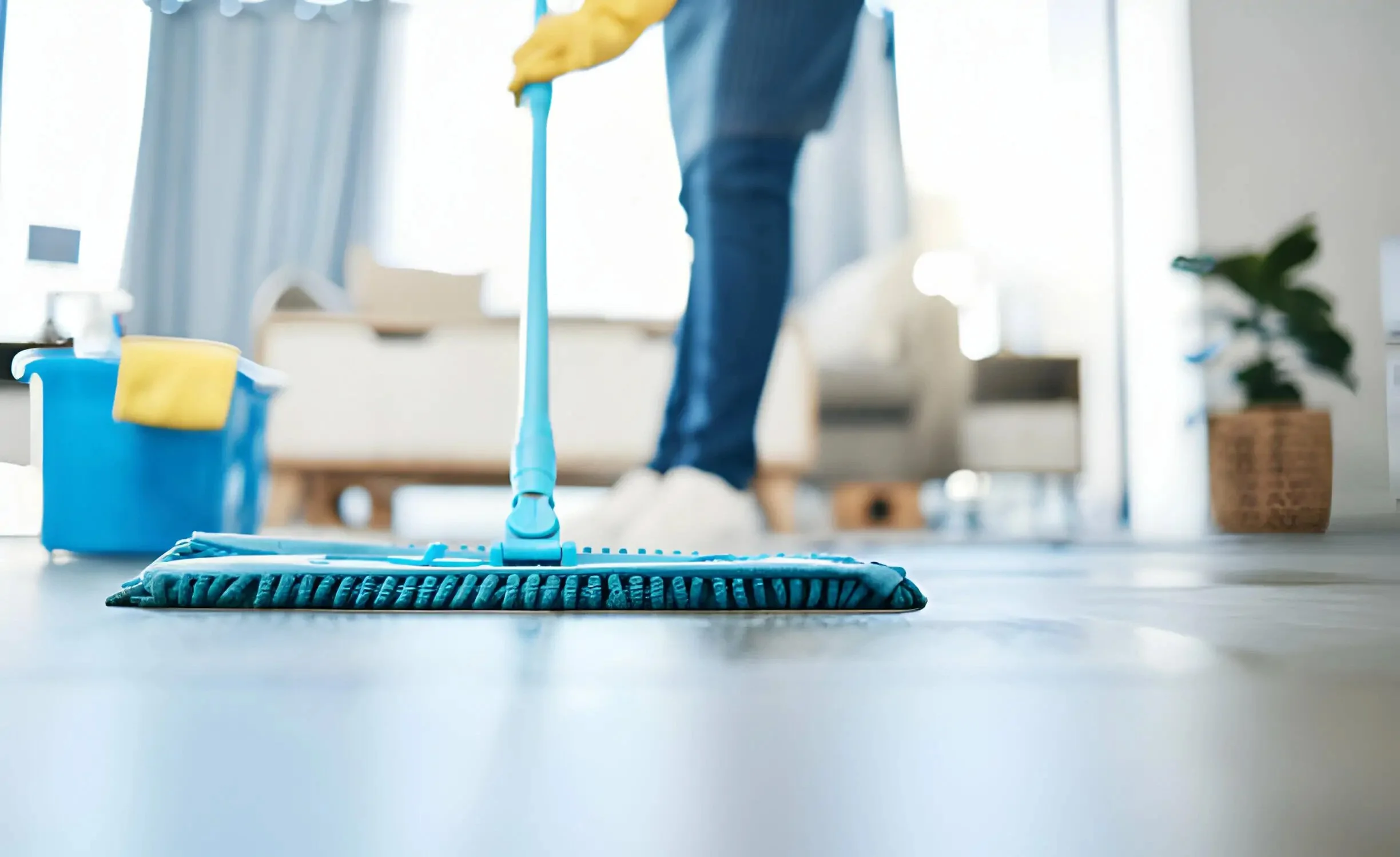 Person mopping floor in a bright living room with blue curtains, potted plant, and a couch in the background.