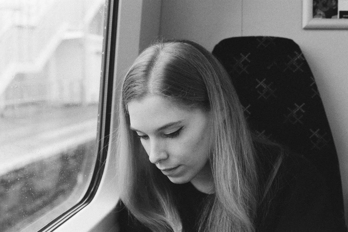 A young person with long hair sitting on a train, looking down, with a window beside them showing a blurred outdoor scene.