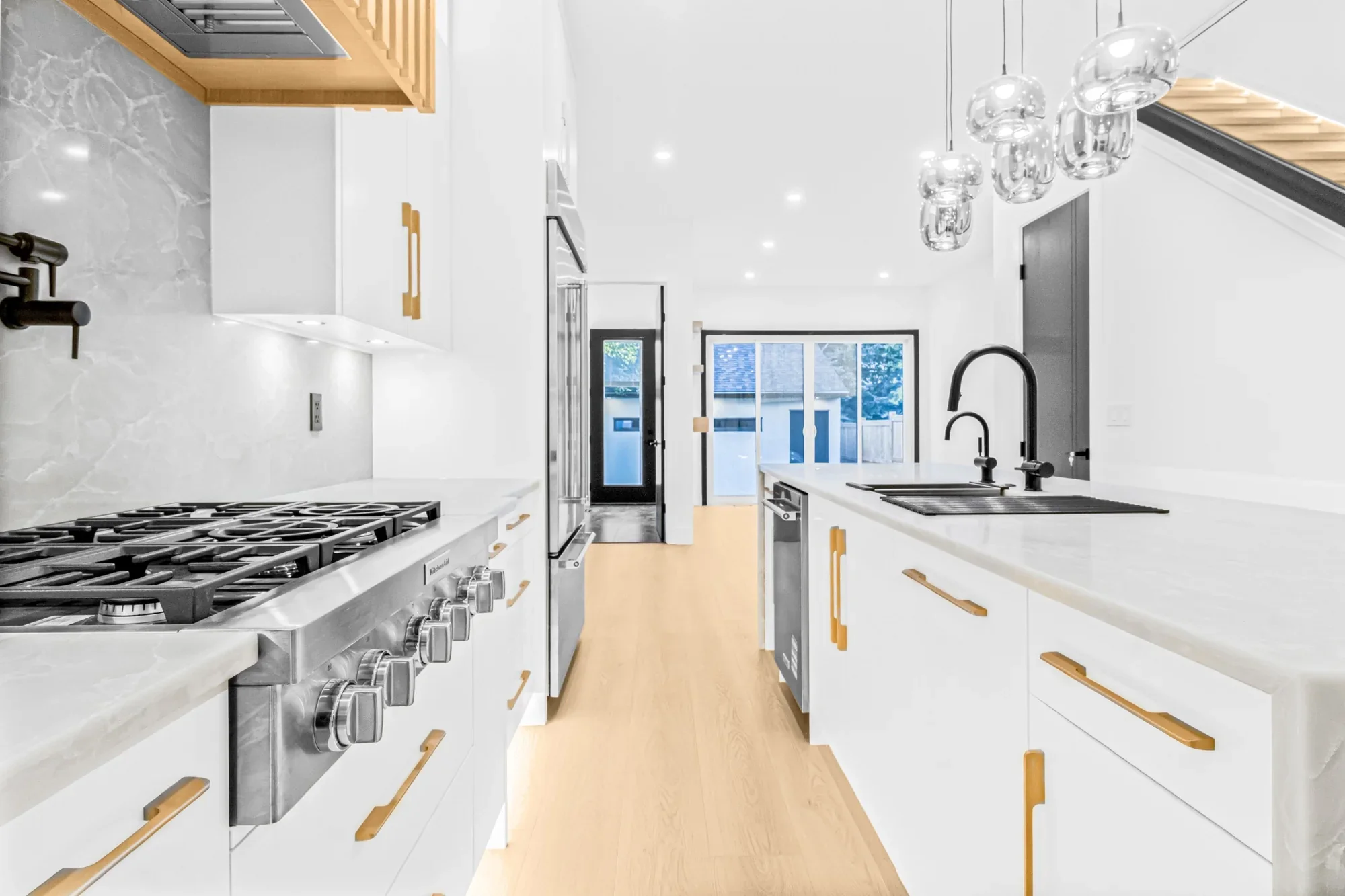 High-end kitchen renovation featuring a waterfall island with integrated sink, professional gas cooktop, custom white cabinetry with brass hardware, pendant lighting, and wood flooring.