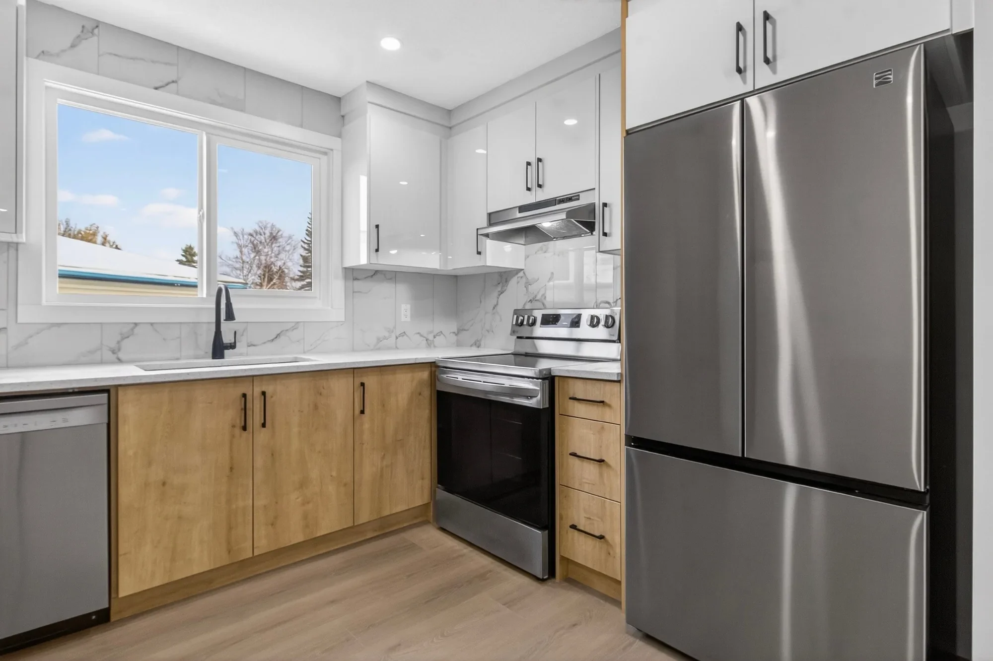 Close-up view of a modern basement kitchen renovation featuring white upper cabinets, wood-tone lower cabinets, marble-style backsplash, stainless steel appliances, and a black kitchen faucet.