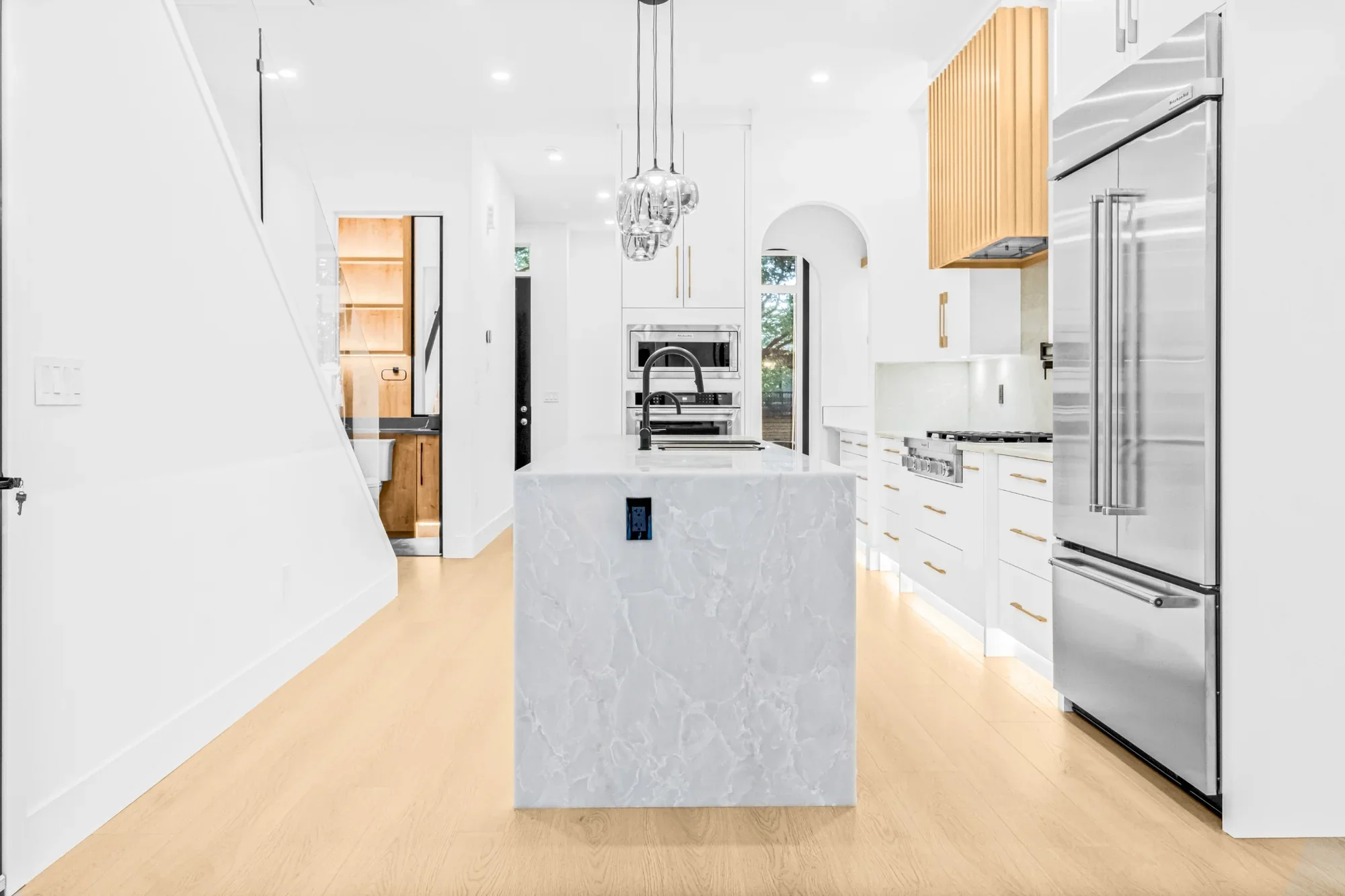 Bright modern kitchen renovation featuring a large waterfall stone island, custom white cabinetry with gold hardware, stainless steel appliances, and warm wood flooring.