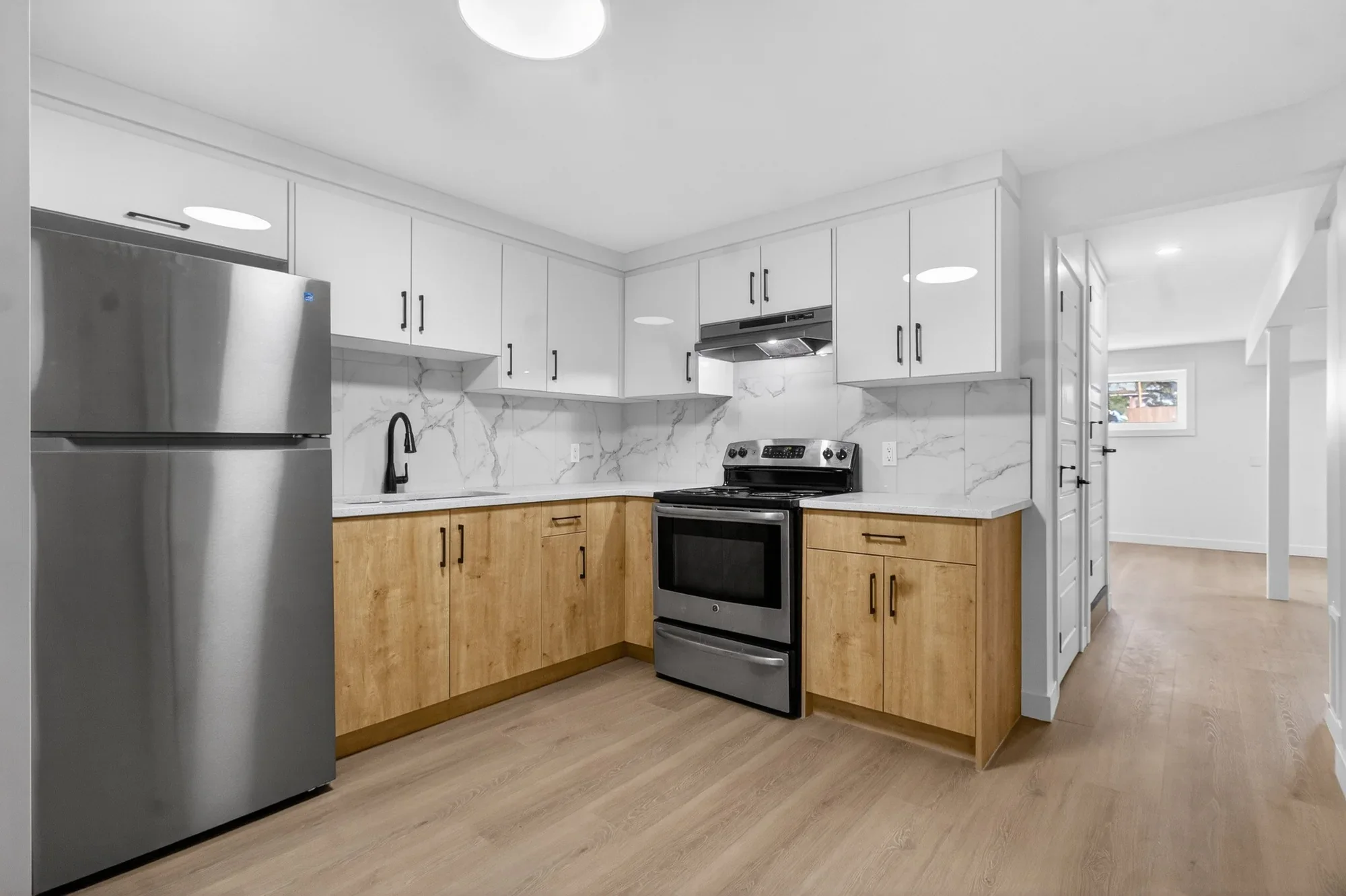 Basement kitchen renovation featuring modern white upper cabinets, wood-tone lower cabinets, marble-style backsplash, stainless steel appliances, and durable wood flooring.