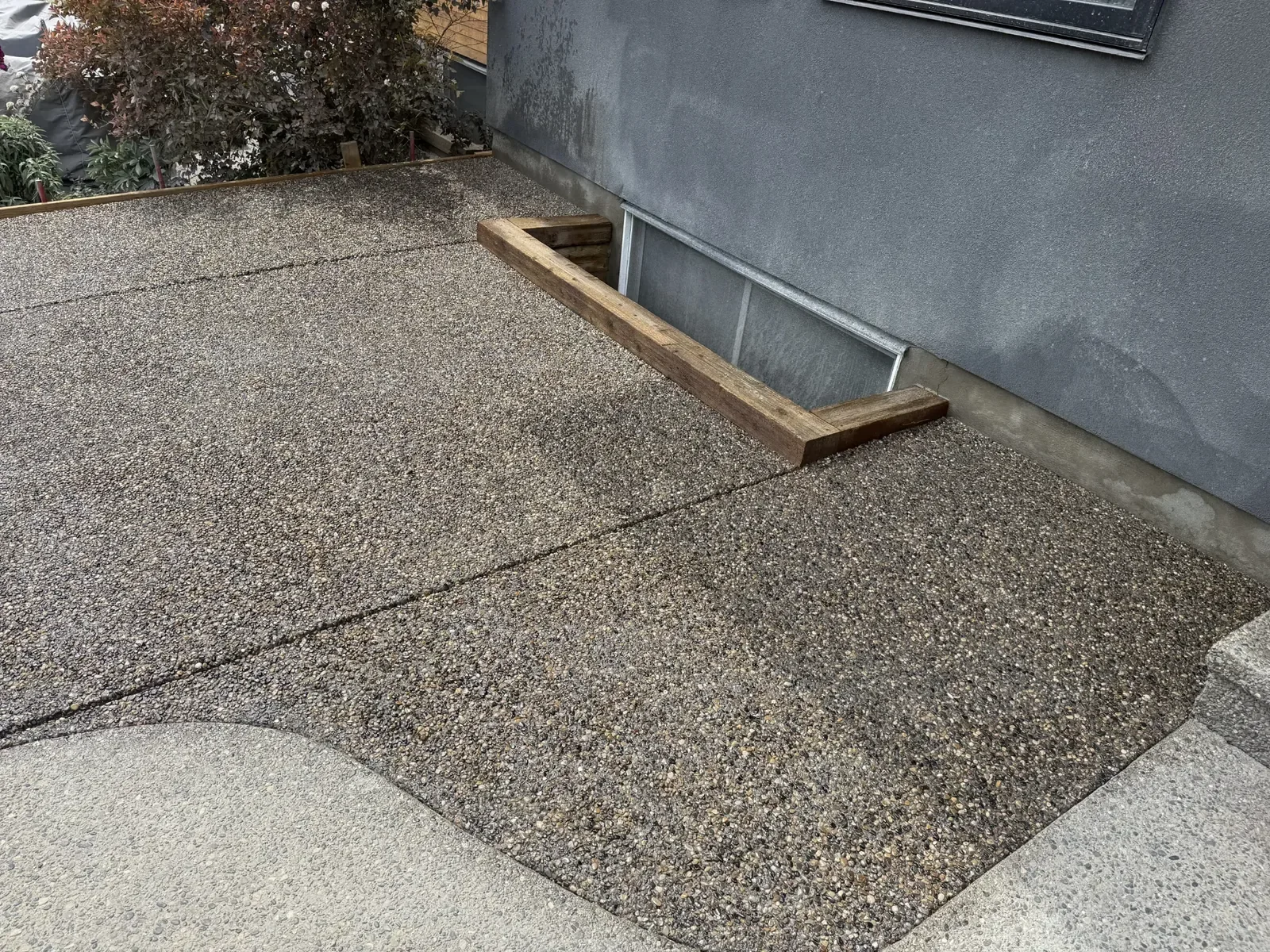 Exposed aggregate concrete patio poured with clean control joints and custom window well framing, providing a durable, slip-resistant surface that integrates seamlessly with the home’s foundation.