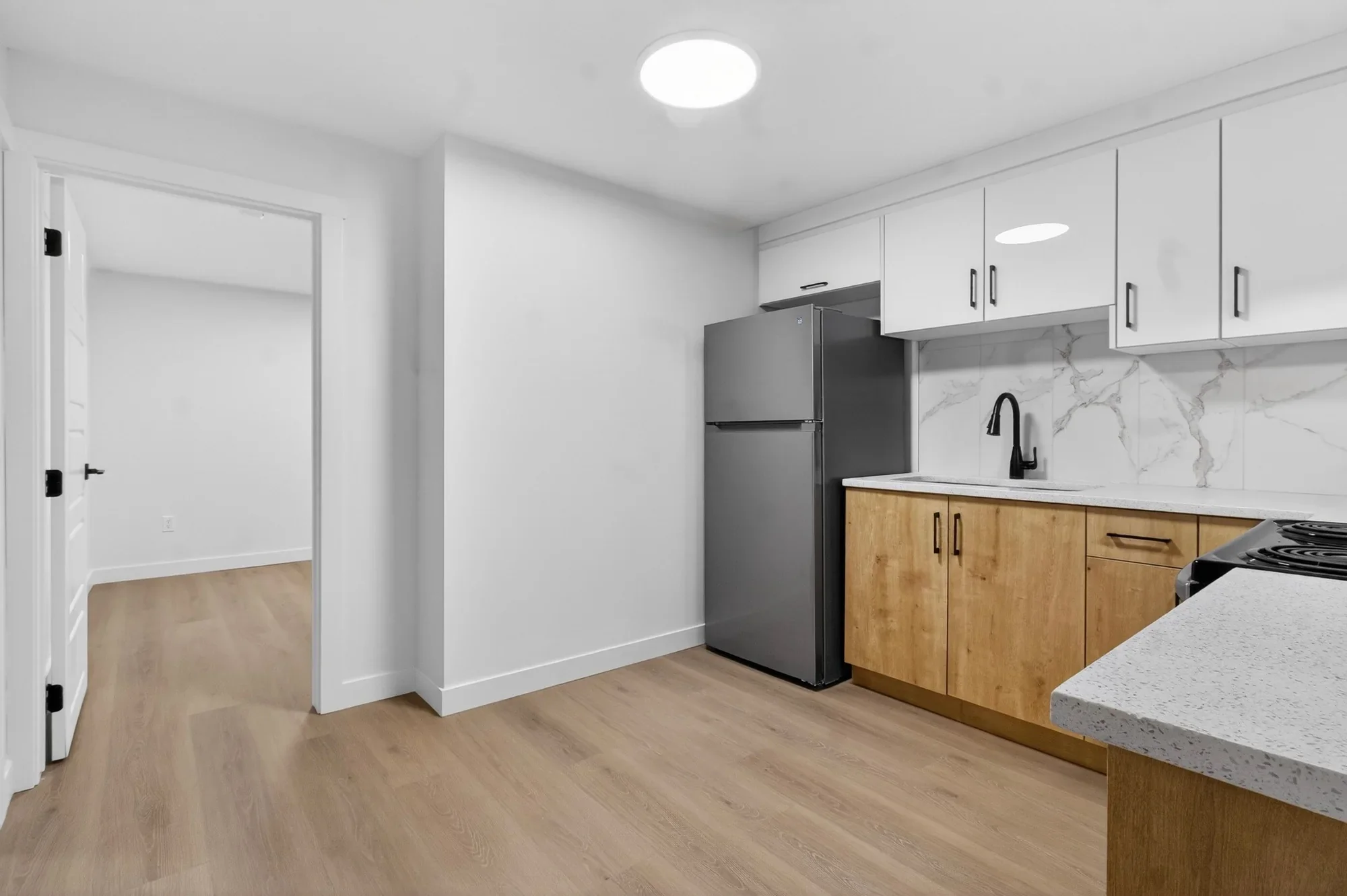 Wide-angle view of a basement kitchen renovation featuring white upper cabinets, wood-tone lower cabinets, quartz countertops, stainless steel refrigerator, marble-style backsplash, and luxury vinyl plank flooring.