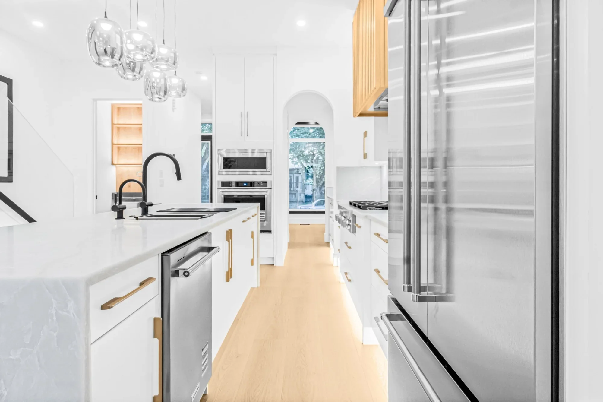 Modern galley-style kitchen renovation with a waterfall island, black fixtures, stainless steel appliances, custom white cabinetry, and warm wood flooring.
