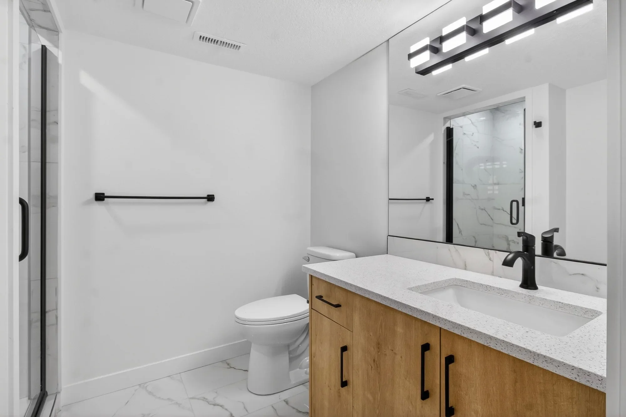 Contemporary bathroom renovation featuring a custom wood-tone vanity, quartz countertop, matte black fixtures, walk-in glass shower, and marble-style tile flooring.