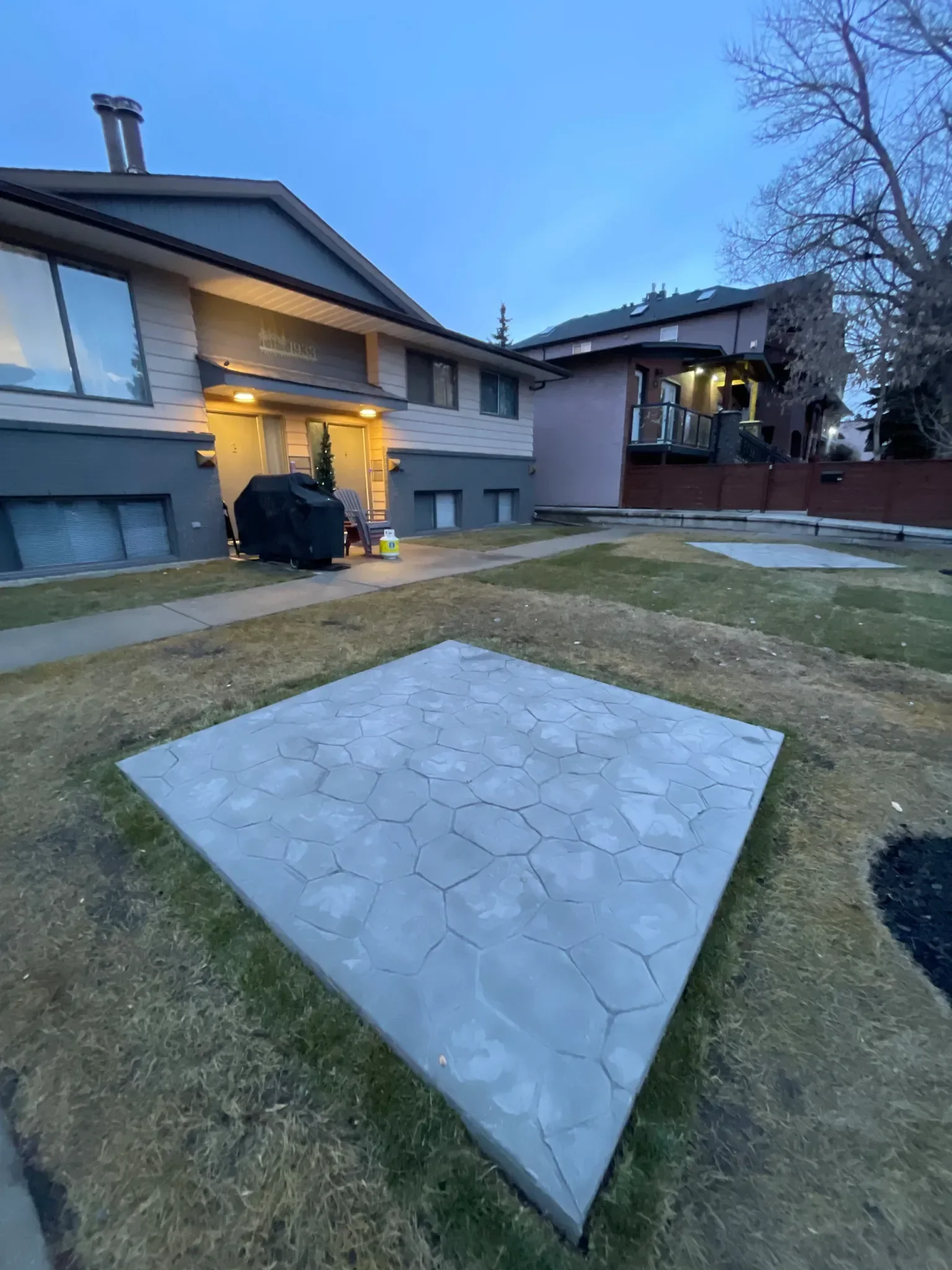 Stamped concrete front yard patio featuring a decorative stone-pattern finish, photographed in evening light to highlight texture and curb appeal. This durable, low-maintenance concrete patio provides a clean outdoor space well-suited for Calgary res