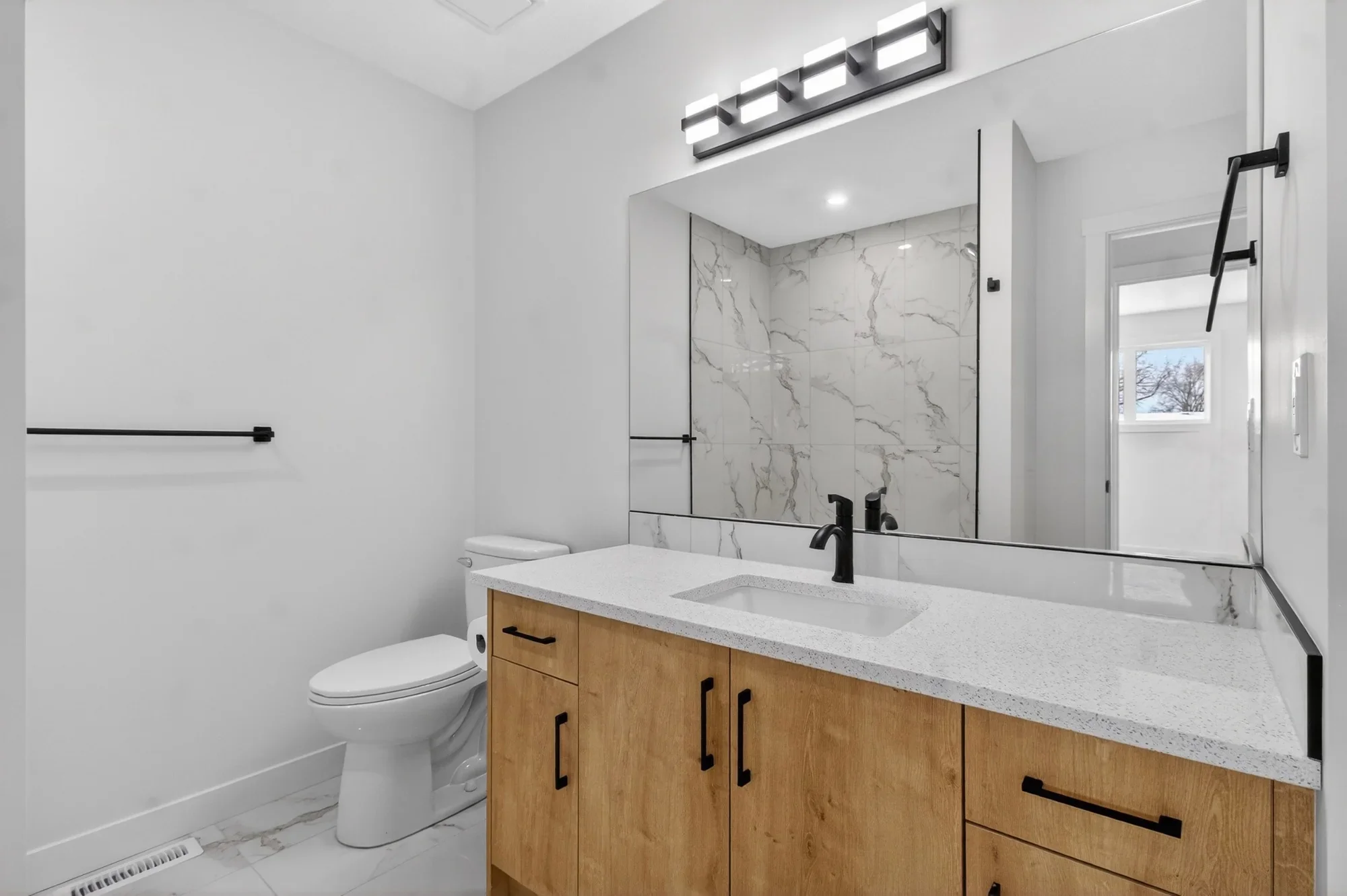 Modern bathroom renovation featuring a custom wood-tone vanity, quartz countertop, matte black fixtures, large mirror, and marble-style tiled shower walls.