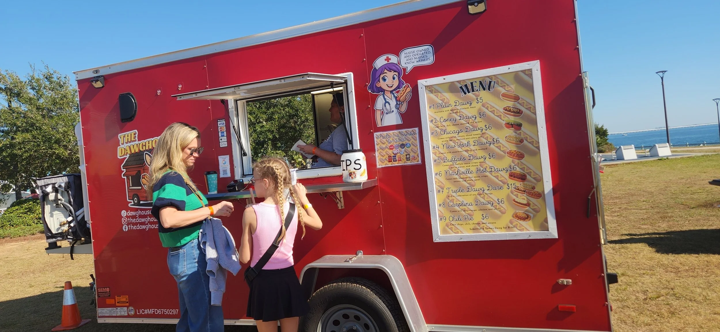 A red food truck with a menu board, serving hot dogs and other fast food, parked next to a grassy area near the water. Two women and a young girl are standing at the window, ordering food, with the ocean and a promenade in the background.
