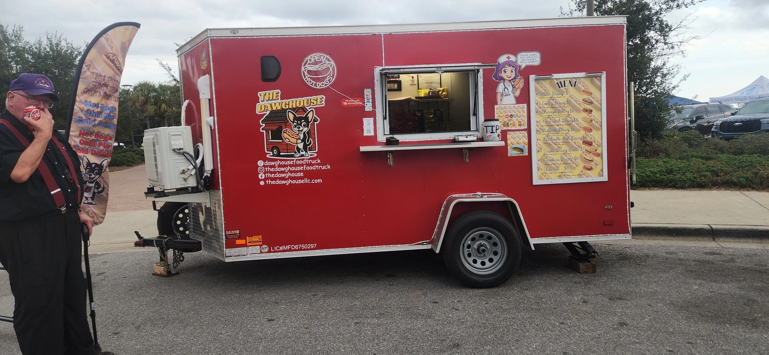 Red food truck named 'The DawgHouse' with a menu panel, cartoon chef, and a man drinking from a Coke cup standing nearby.