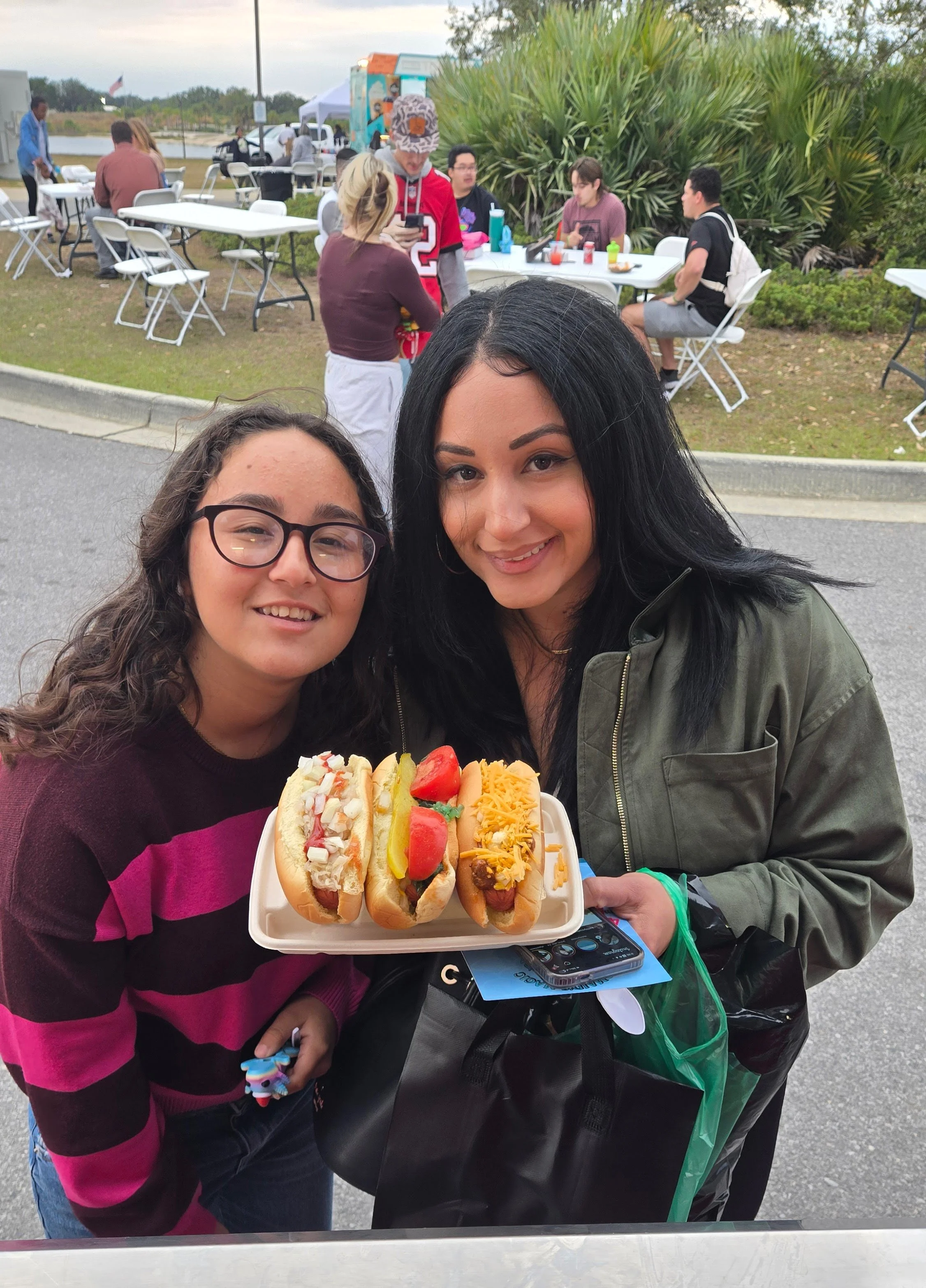 Two women smiling, holding a tray of hot dogs topped with various ingredients at an outdoor event with tables and people in the background.