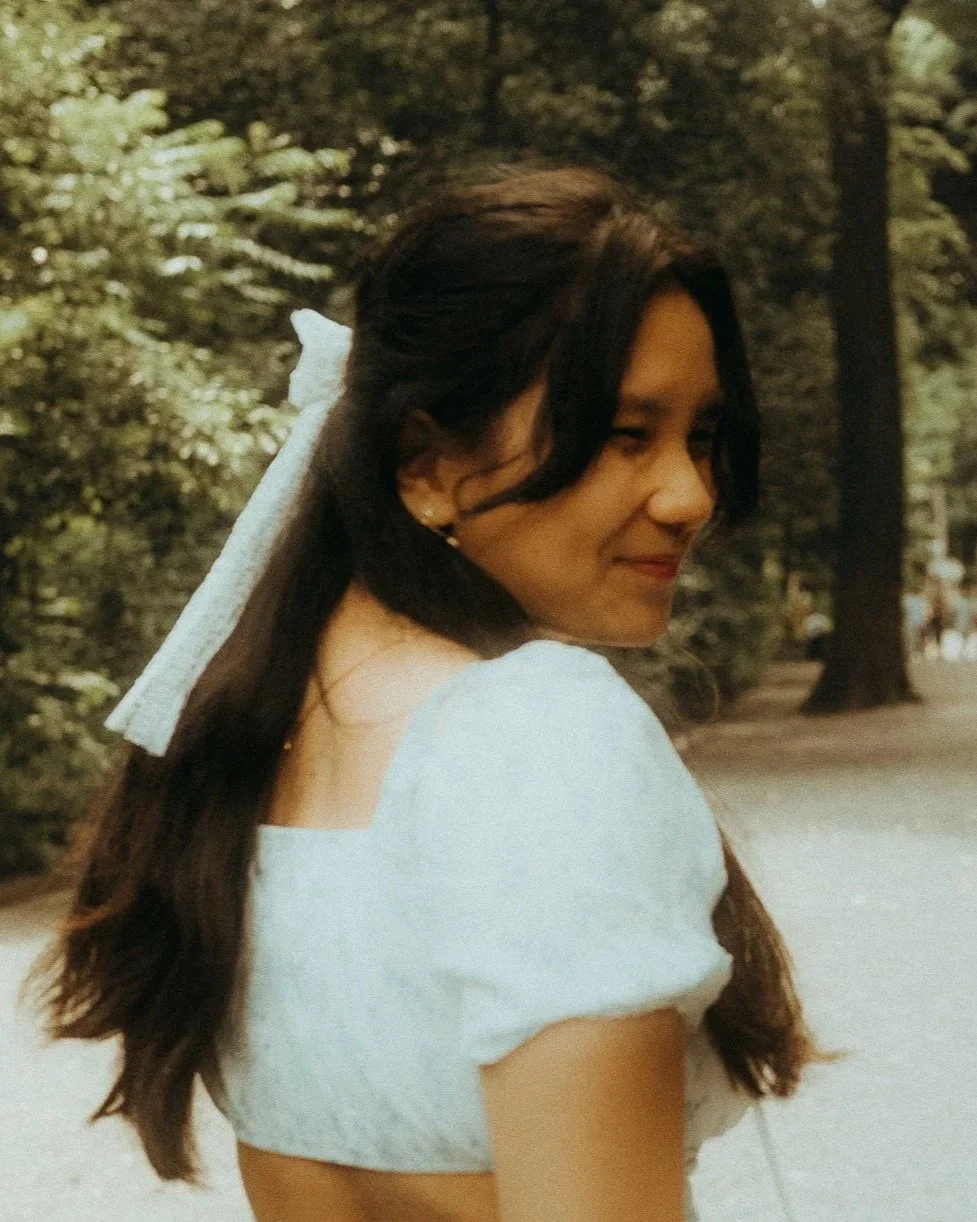A young woman with dark hair wearing a white dress with puffed sleeves, smiling with her eyes closed, in a park with trees and greenery in the background.