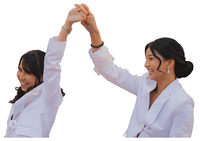 Two women in white suits are smiling and holding hands in a celebratory gesture.