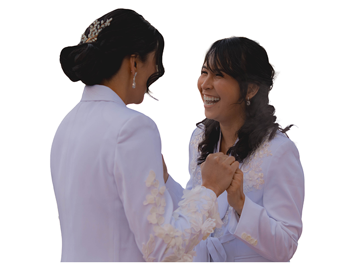 Two women in wedding suits holding hands and smiling at each other.