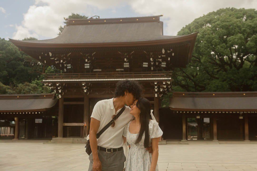 A young couple standing close, smiling, with their foreheads touching in front of a traditional Japanese temple gate surrounded by trees.