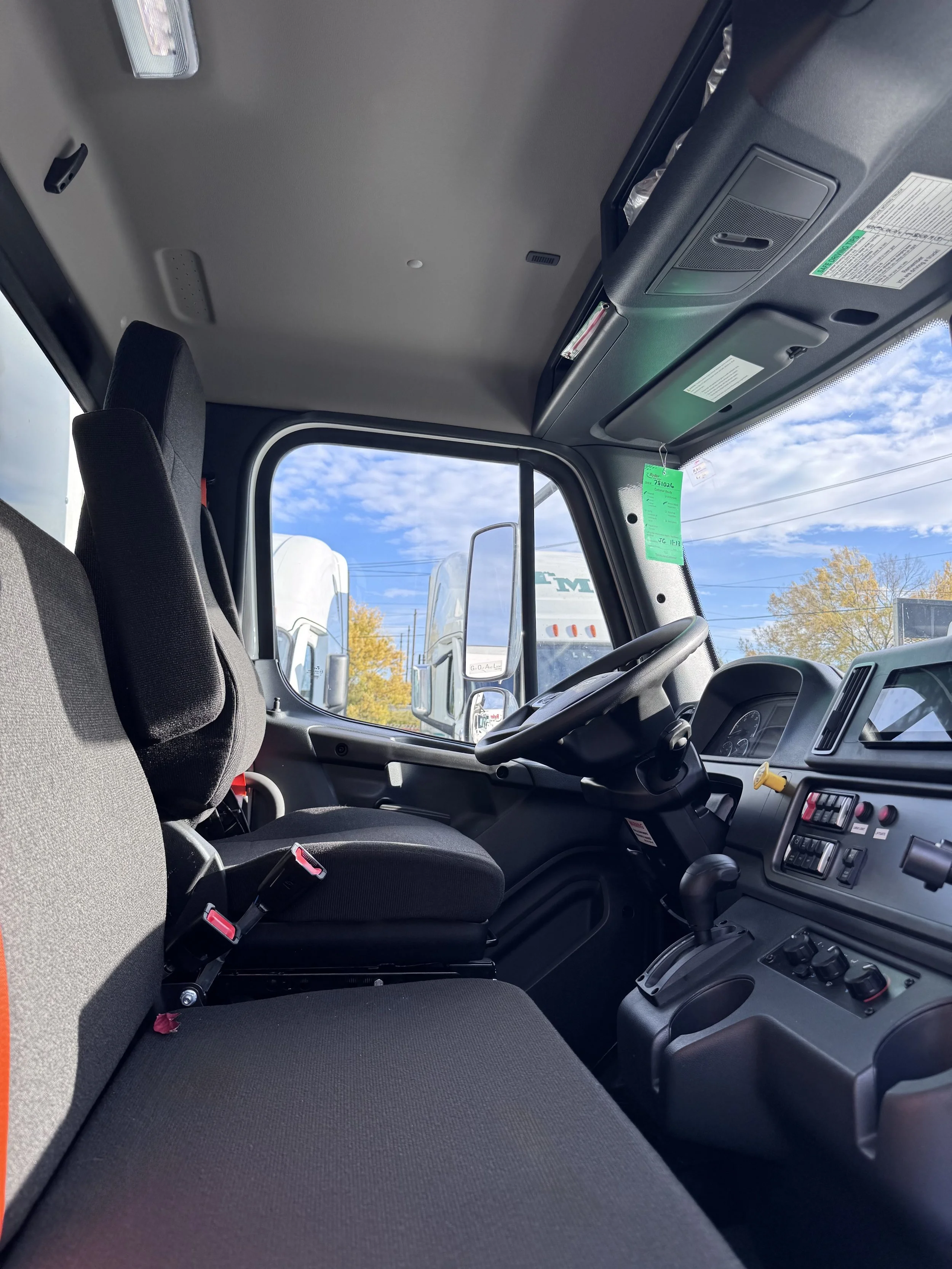 Inside view of a truck cab showing the driver's seat, passenger seat, steering wheel, dashboard, and large front window with a view of multiple trucks and a partly cloudy sky outside.