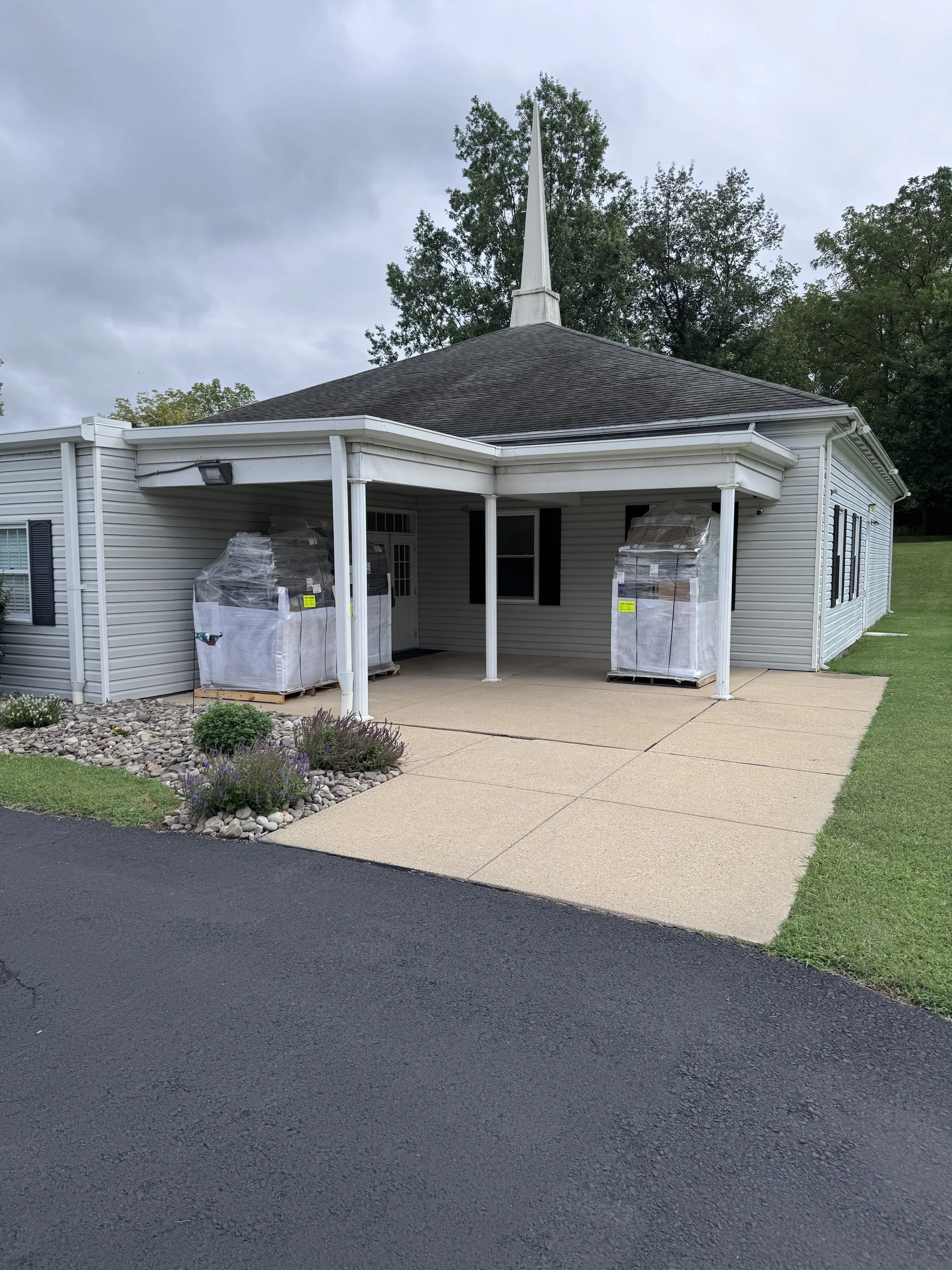 A small church with a steeple, vinyl siding, and a covered porch, with two large warping machines on pallets in plastic covers on the porch.