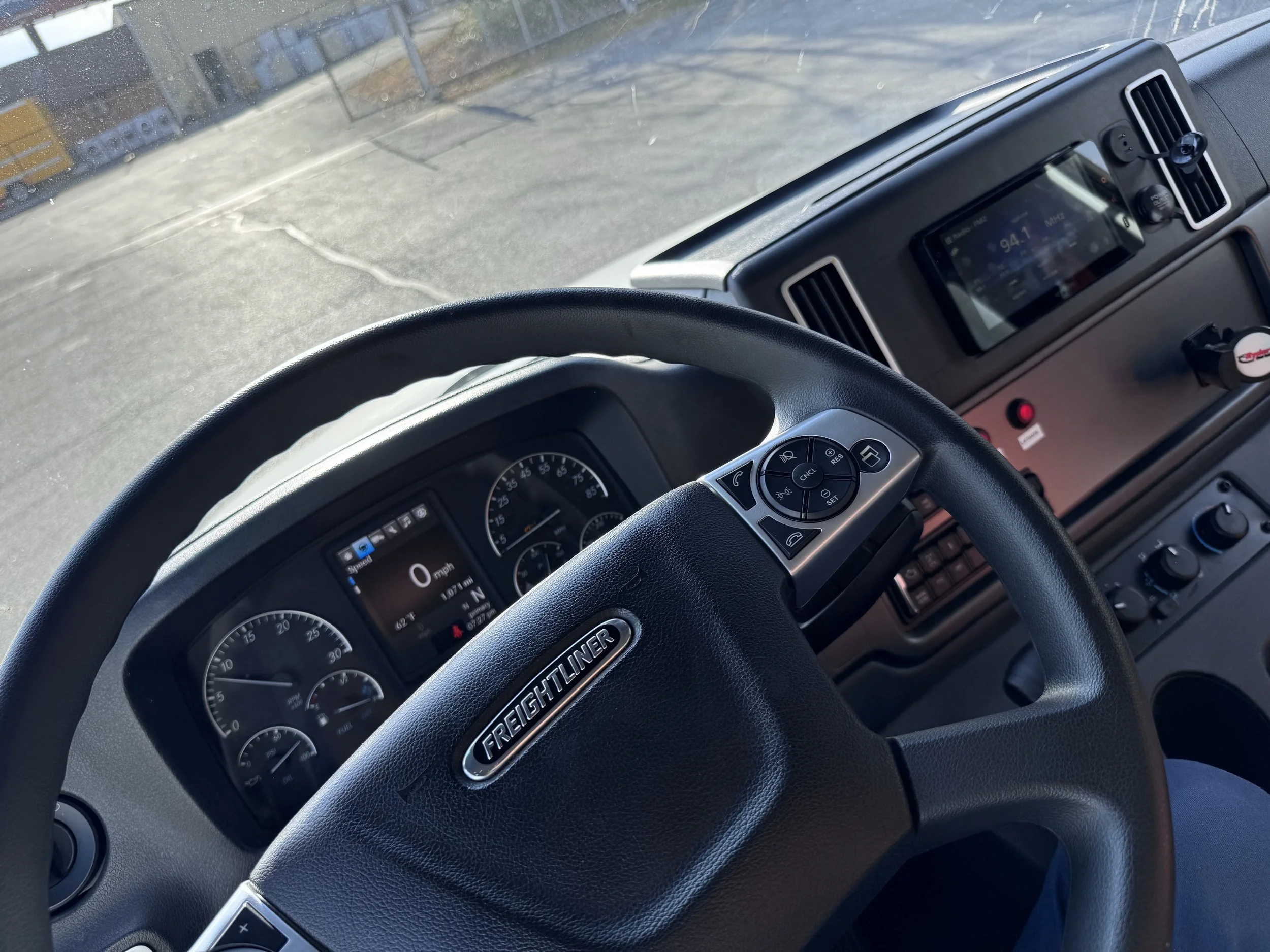 Inside the driver's cabin of a Freightliner truck, showing the steering wheel, dashboard with speedometer and gauges, and a dashboard display showing 0 mph. The background shows an outdoor area with a basketball hoop and a fenced yard.