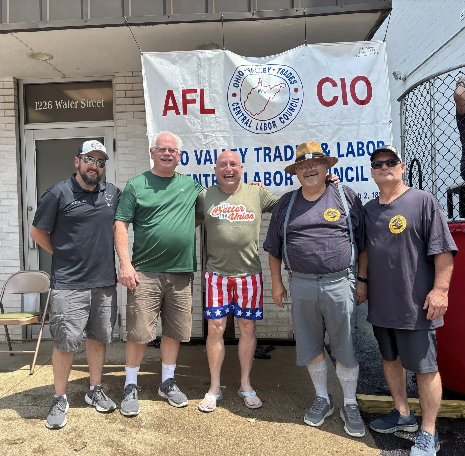 Group of five men standing outdoors in front of a banner for the Ohio Valley Trades and Labor Center Labor Council, with some smiling and casual clothing, including shorts, t-shirts, and hats.