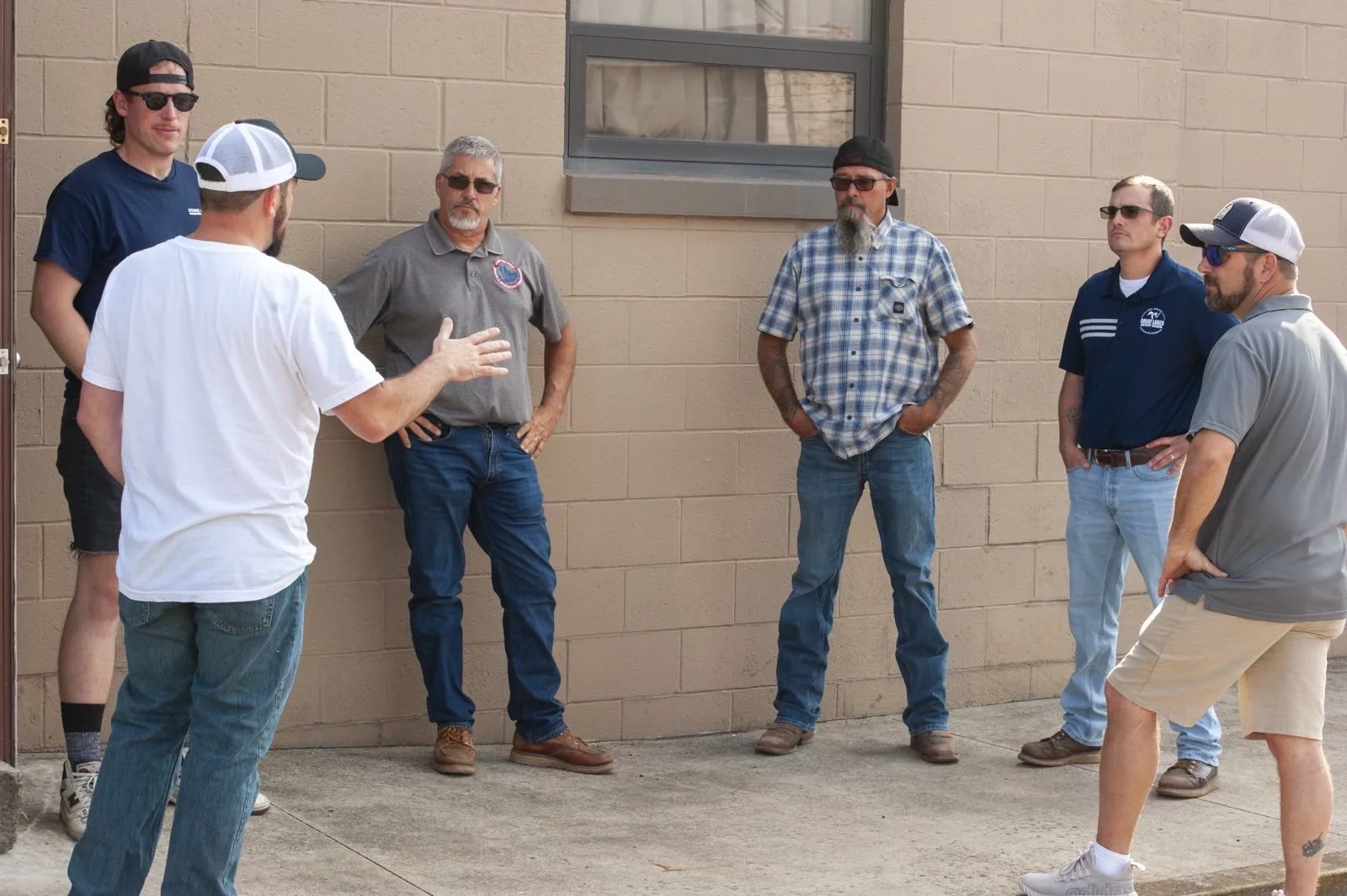 Six men standing and listening to a man in a white t-shirt speaking. They are outdoors, against a brick wall with a window.