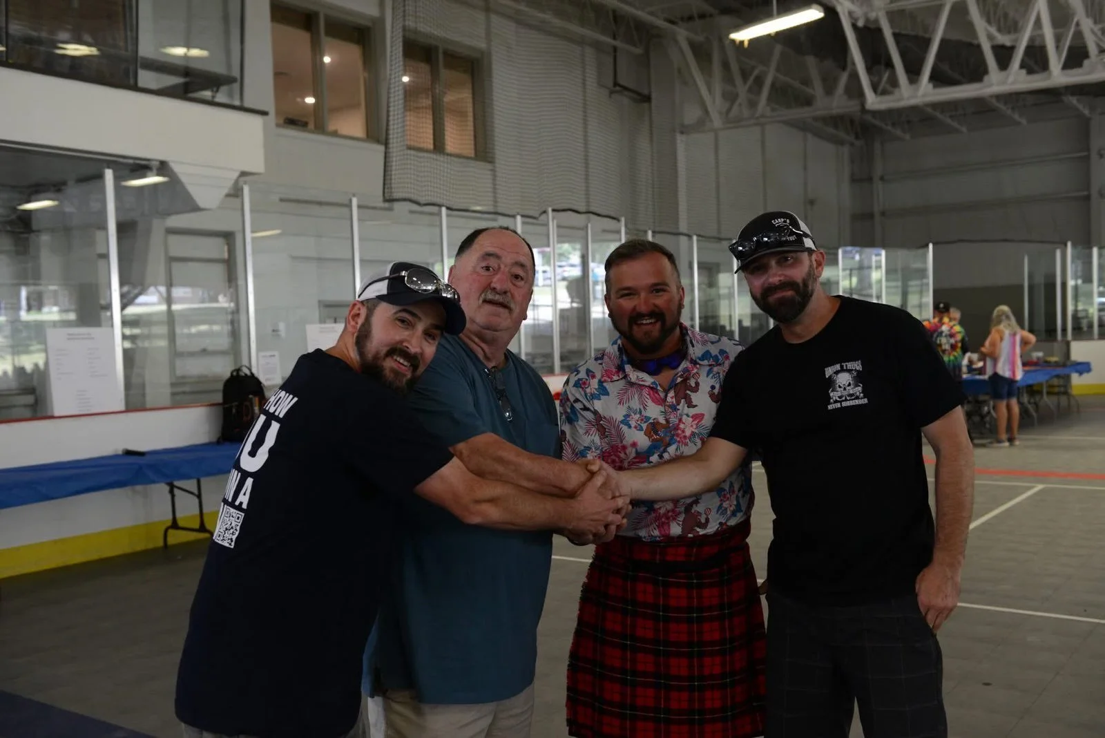 Four men shake hands in an indoor ice rink, with two in dark shirts wearing hats, one in a blue shirt, and one in a colorful Hawaiian shirt and red plaid kilt, smiling at the camera.