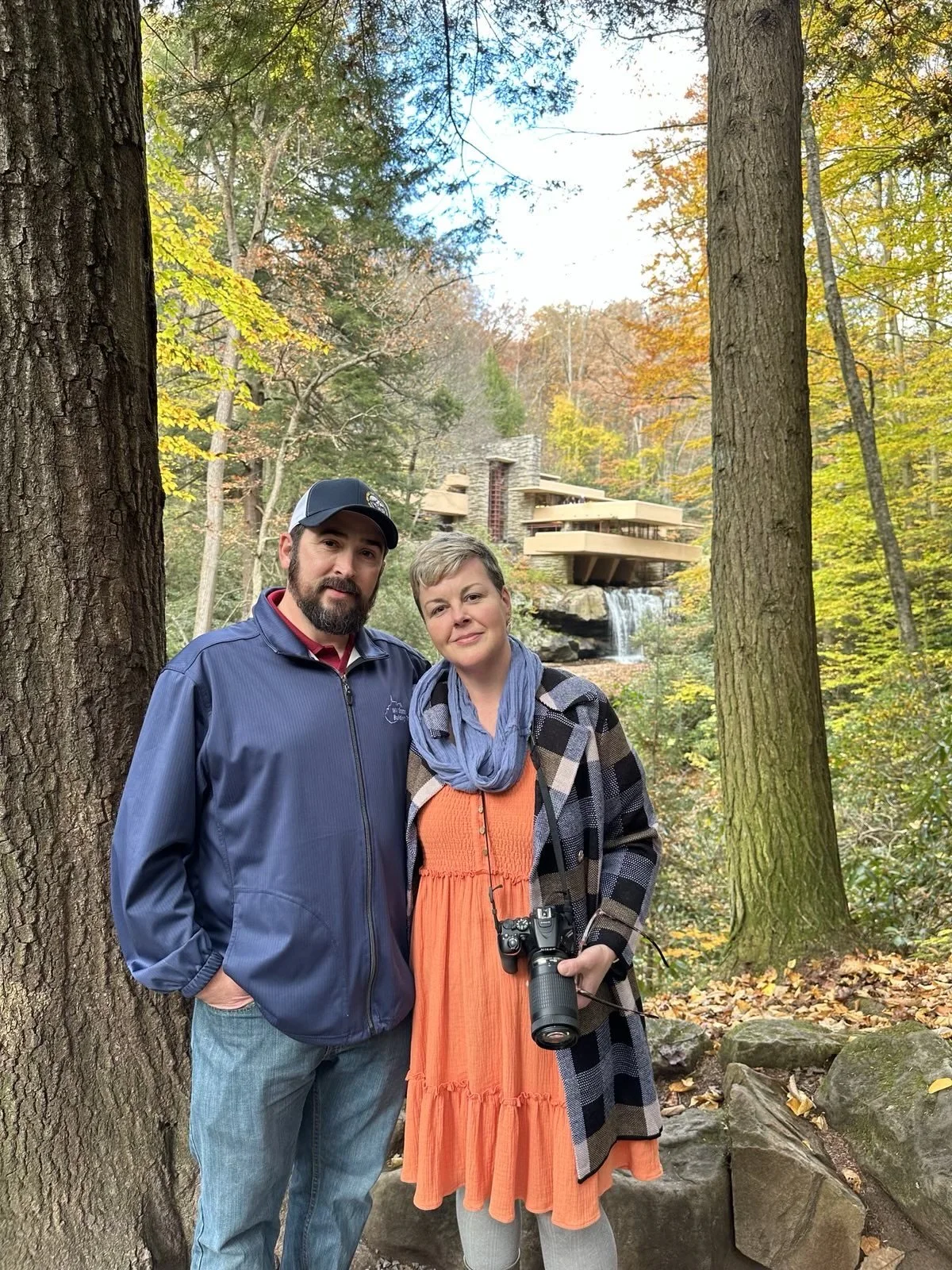 A man and woman standing close together outdoors in a forested area with trees and autumn foliage, behind them a modern house with a waterfall feature.