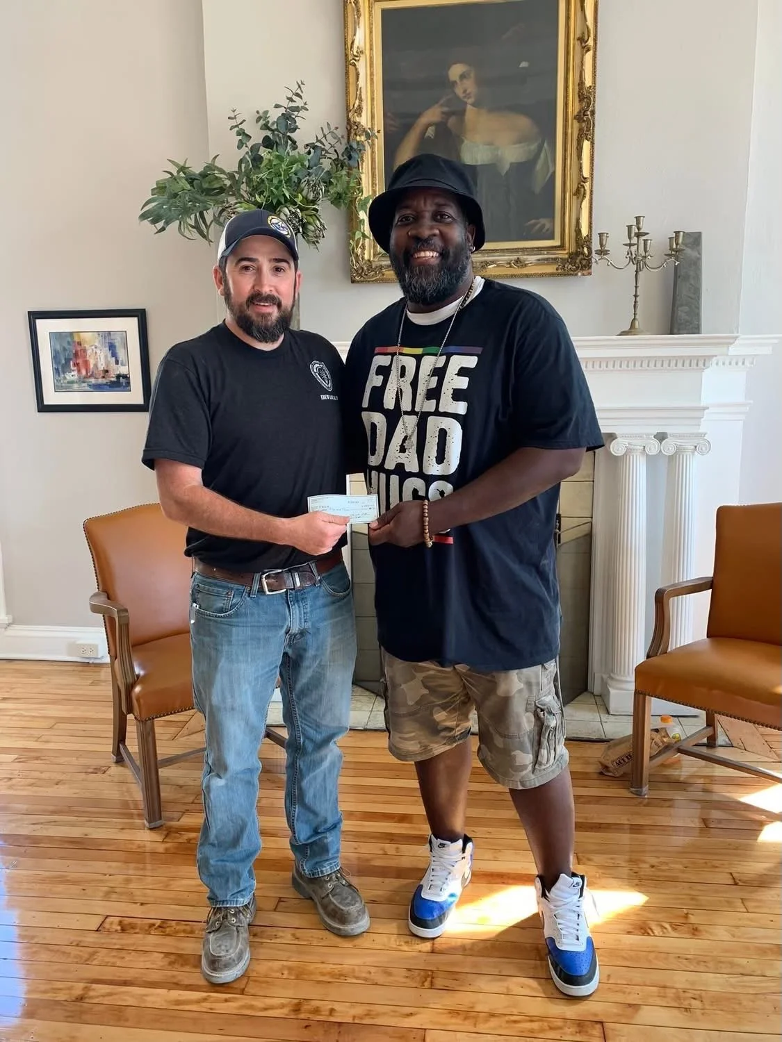 Two men standing indoors, smiling, exchanging a check. One is wearing a black T-shirt, jeans, and a cap; the other is wearing a black T-shirt with the text 'FREE DAD HUGS,' camouflage shorts, and sneakers. There is a painting of a woman on the wall behind them, a fireplace with decorative items on the mantel, and two yellow chairs placed against the walls.