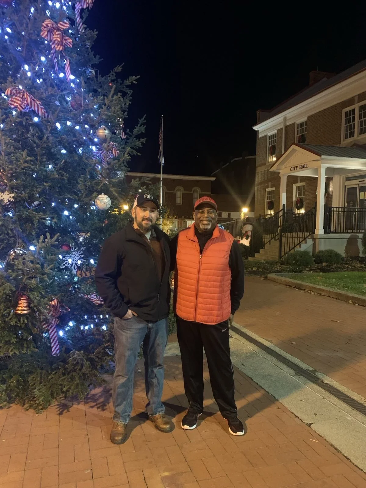 Two men standing next to a decorated Christmas tree in front of a city hall building at night.