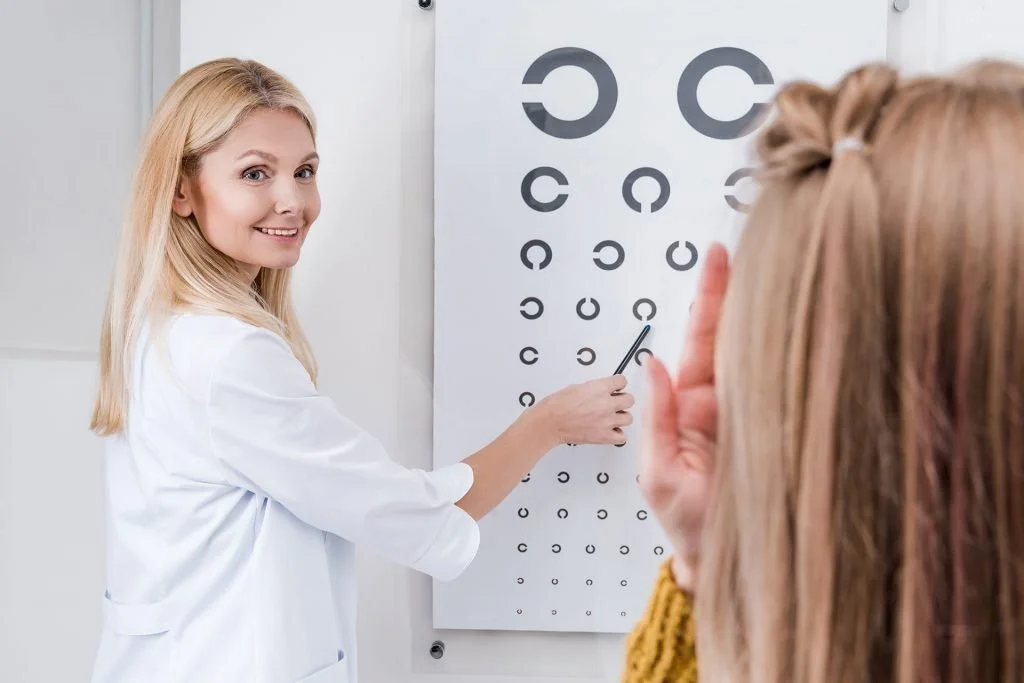 Optometrist in white coat demonstrating eye chart to a patient in an eye examination room.