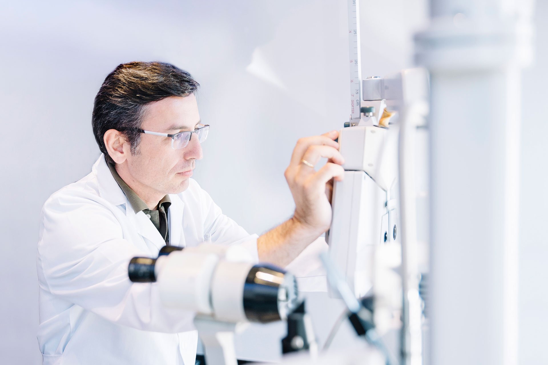 A male scientist or doctor wearing glasses and a white lab coat working with medical equipment or machinery in a laboratory setting.