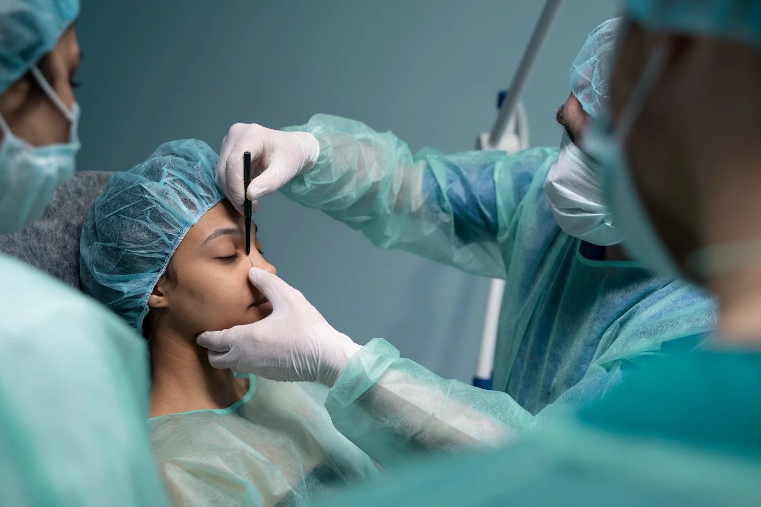 Medical professionals performing a cosmetic procedure on a female patient. The patient is lying down with her eyes closed, wearing a surgical cap. One medical professional is holding her chin with one hand and using a tool on her forehead with the other, while another assists.