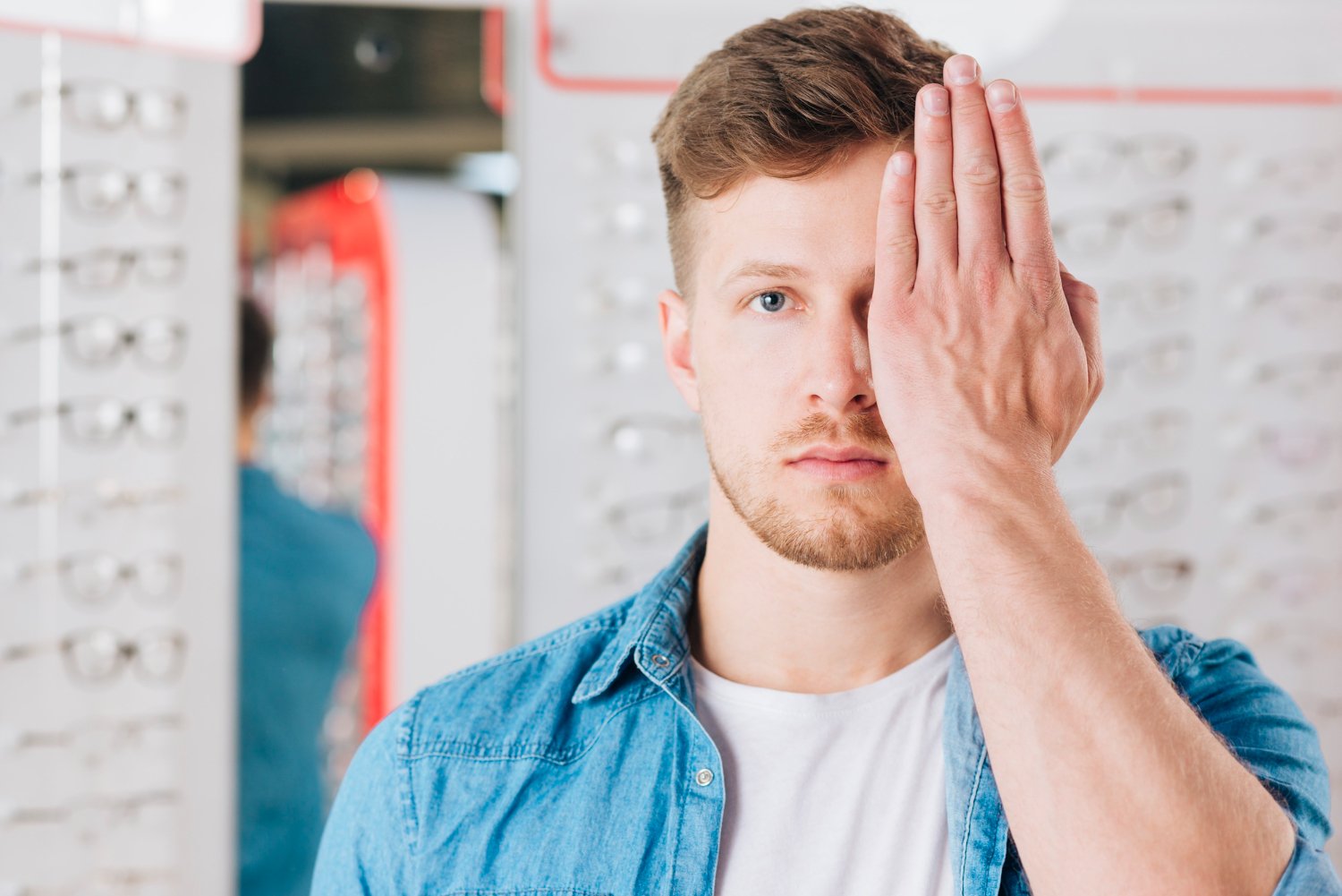 A young man with a beard and short hair wearing a denim shirt and white t-shirt, holding his hand against one side of his face, standing in front of mirrors in an eyewear store.