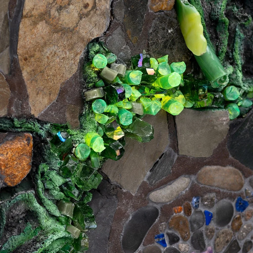 Close-up view of a mosaic wall with green glass tiles and stones of different colors and shapes.