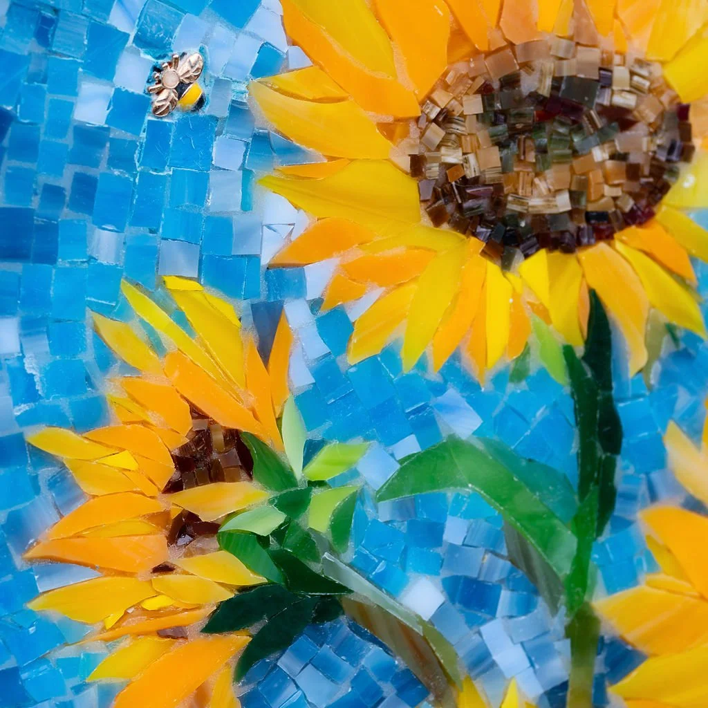 Mosaic artwork depicting a sunflower with yellow petals, green leaves, and a brown center, set against a blue sky background.